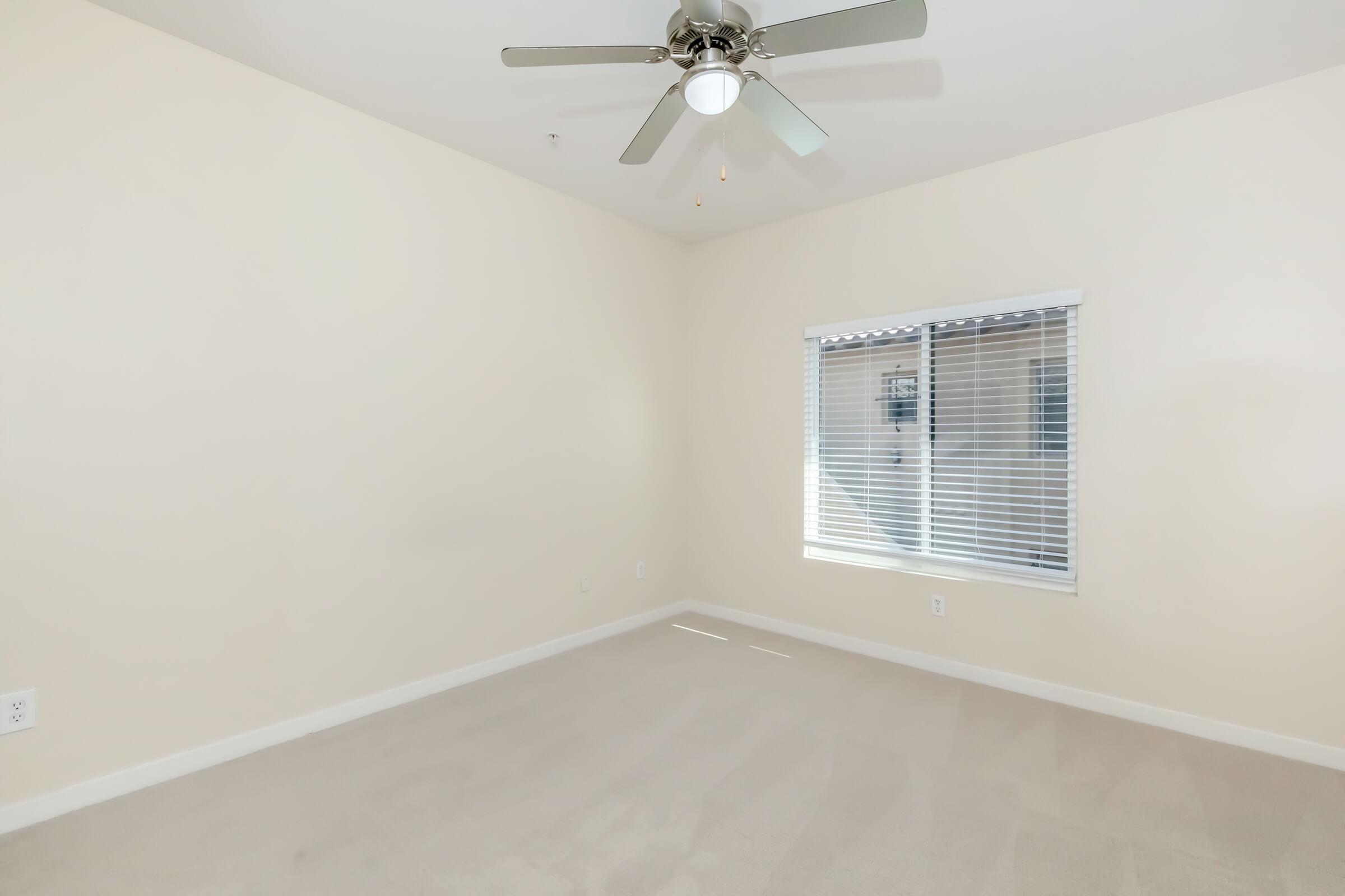 An empty, well-lit room with light beige walls and carpeting. A ceiling fan is installed, and there is a window with white blinds allowing natural light to enter. The space is uncluttered, suggesting it could be a bedroom or a home office.