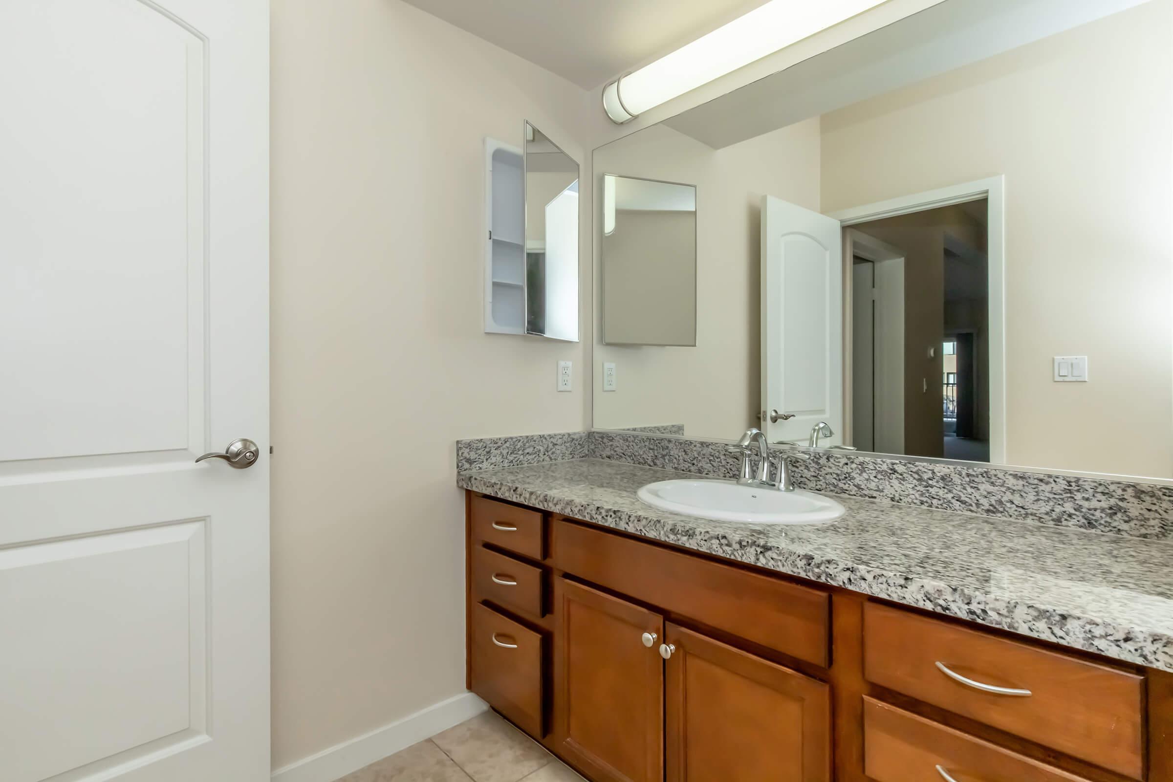 Bright and modern bathroom featuring a granite countertop with a sink, wooden cabinetry, a large mirror, and neutral wall colors. The space is well-lit with overhead lighting and includes a door leading to another room. The floor is tiled, adding to the contemporary aesthetic.