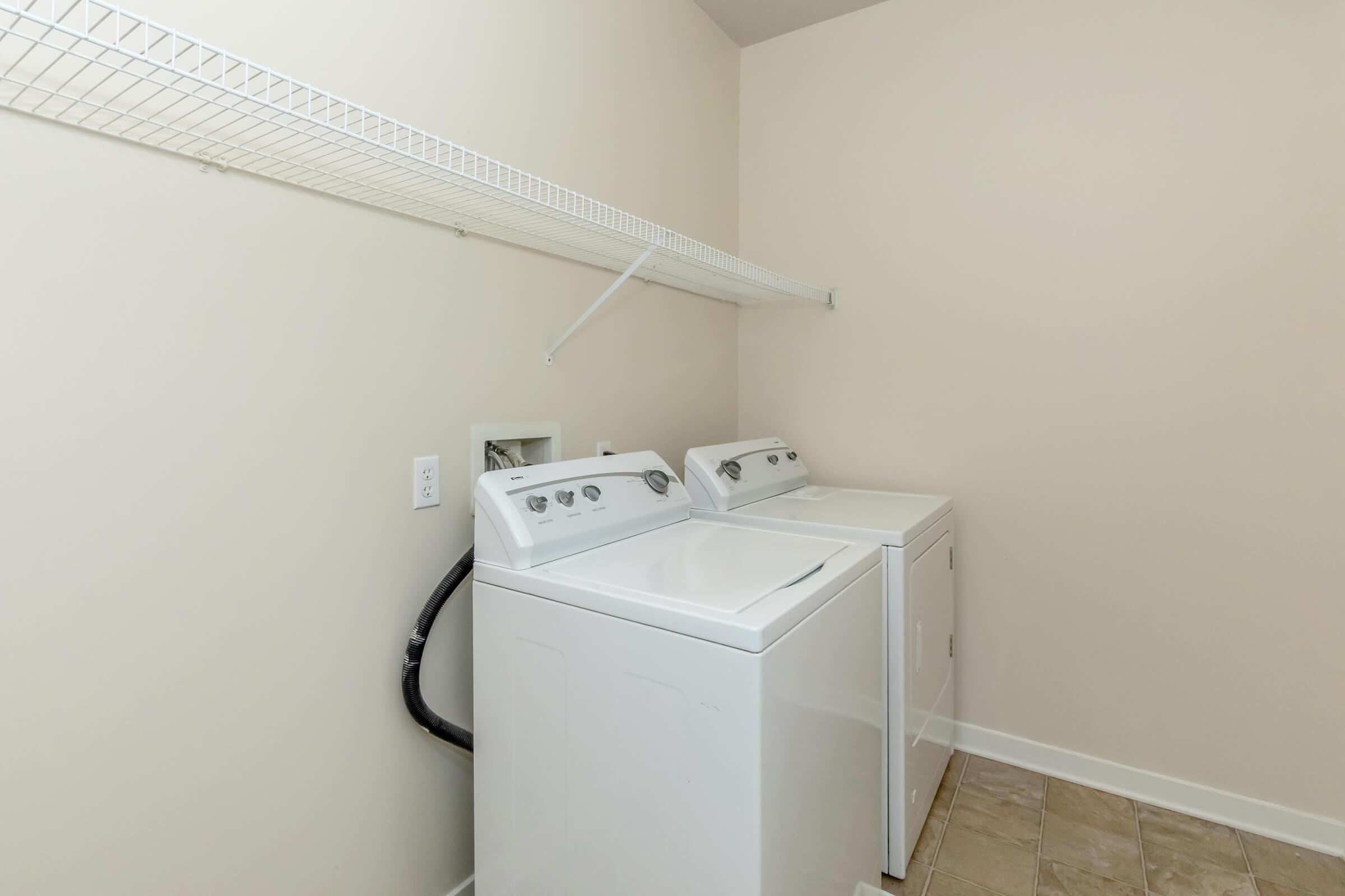 Two white washing machines side by side in a clean, light-colored laundry room. A wire shelf is mounted above them, and the floor is tiled. The walls are plain and beige, creating a minimalist and organized space.