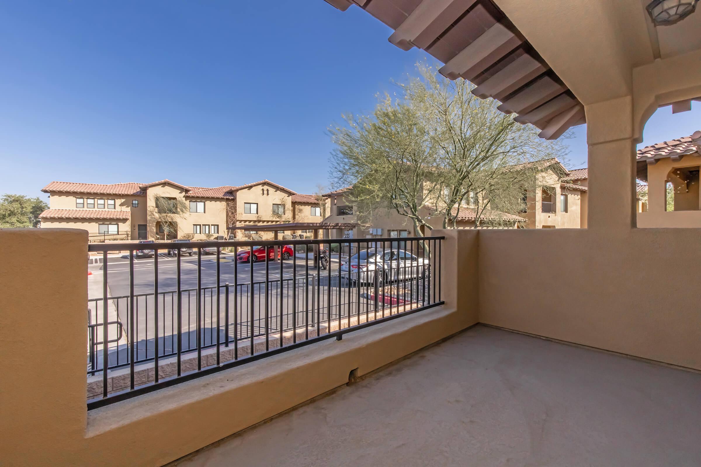A view from a balcony overlooking a parking area and residential buildings under a clear blue sky. The setting features tan-colored walls and nearby trees, creating a peaceful ambiance.
