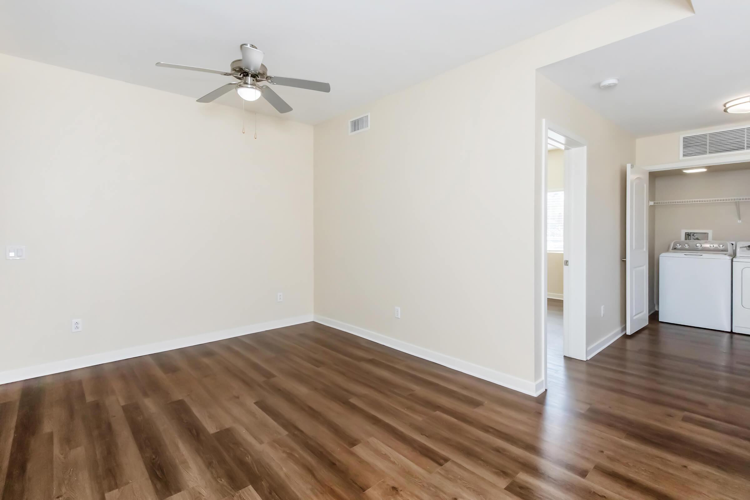 A spacious, well-lit room with light beige walls and hardwood flooring. A ceiling fan is mounted above. On one side, there is an entrance to a laundry area with appliances visible. The room is empty and ready for furniture arrangement. Natural light is coming from a nearby window.