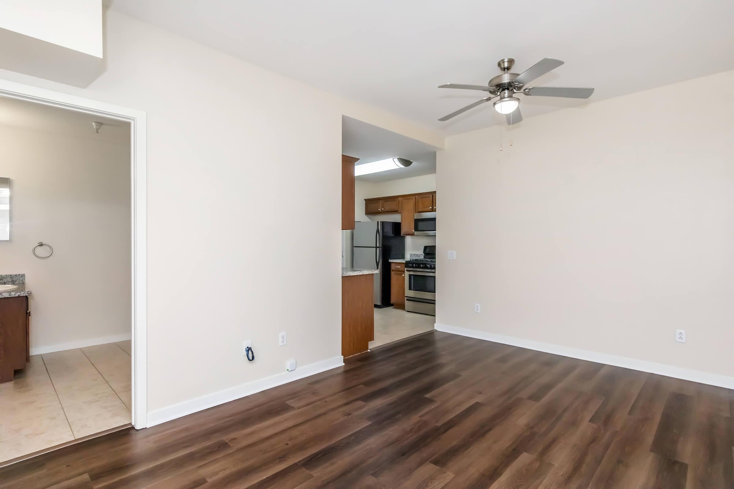 A spacious living area featuring light-colored walls, a ceiling fan, and hardwood-style flooring. To the left, there's an entry to a bathroom with a sink, while the adjacent kitchen is visible with wooden cabinetry and modern appliances. Natural light brightens the room, creating an inviting atmosphere.