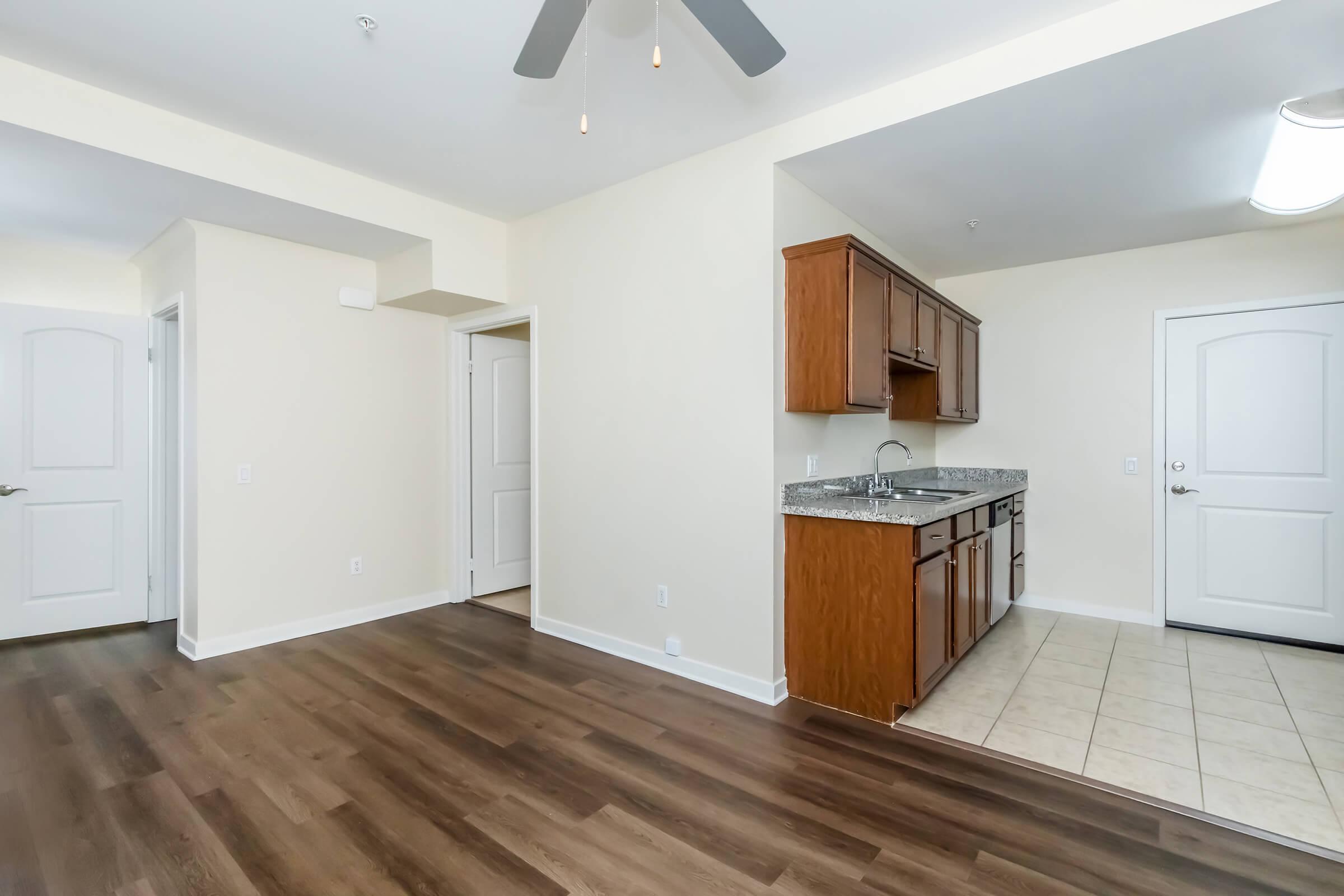An interior view of a modern apartment showing a spacious living area with wood-style flooring, a ceiling fan, and a kitchen area with wooden cabinets and granite countertops. The design is open with light-colored walls and a door leading to another room.