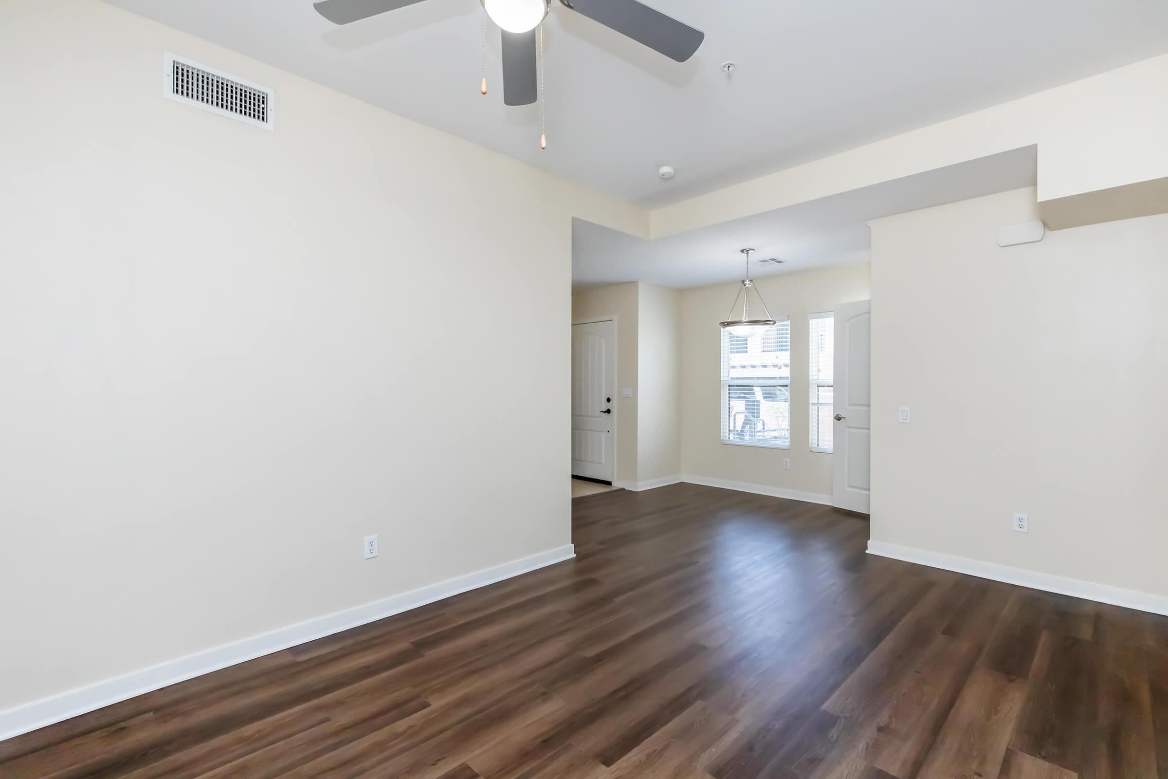 Spacious, empty living area with light-colored walls and brown laminate flooring. A ceiling fan is visible, and there's an adjacent entryway with a door and window. Natural light spills in from the window, creating a bright atmosphere. A pendant light hangs above, adding to the modern feel.