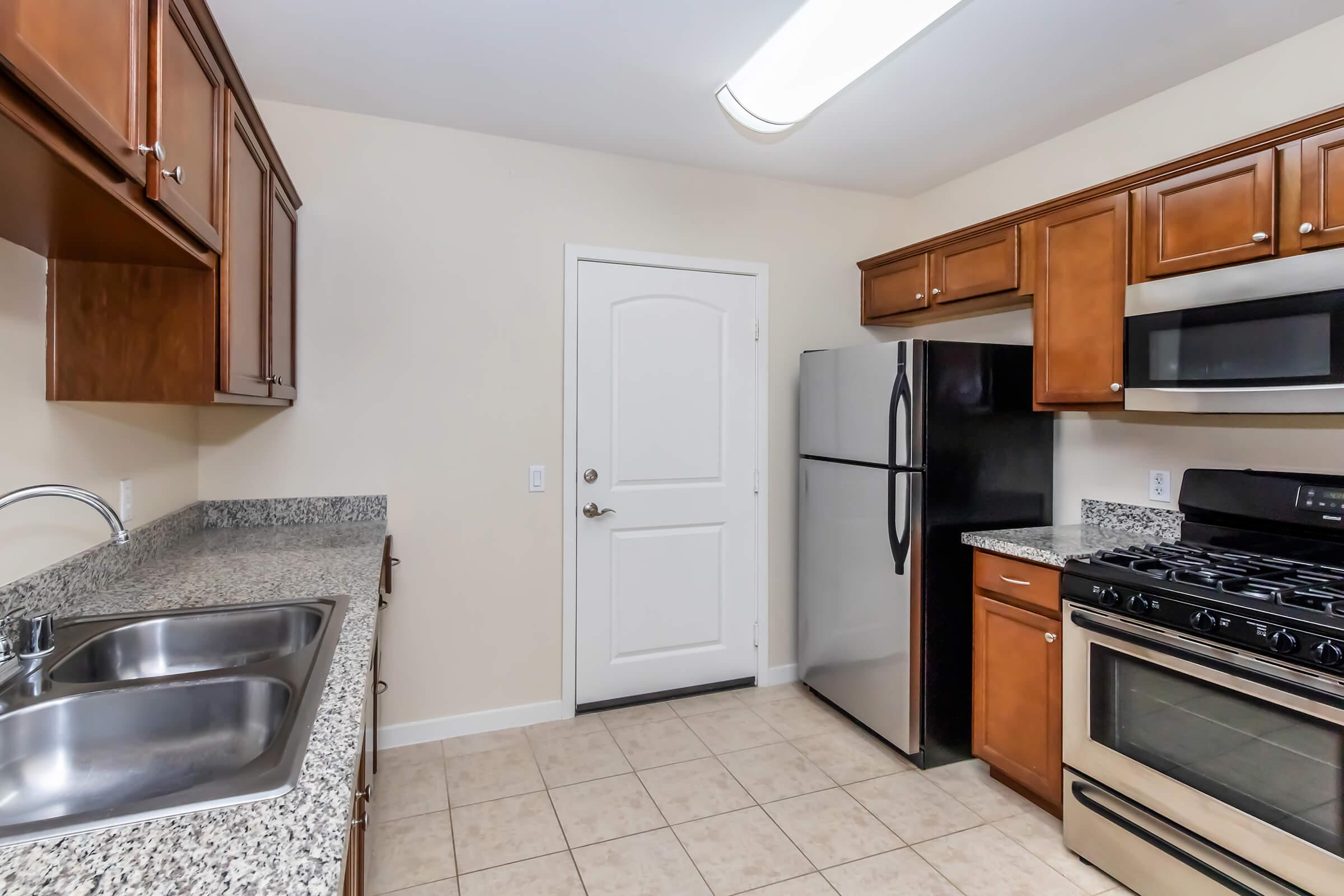 Modern kitchen featuring granite countertops, wooden cabinets, a double sink, and stainless steel appliances including a refrigerator and a gas stove. A door leads outside, and the floor is tiled. The overall design is clean and functional, with an open layout and good lighting.