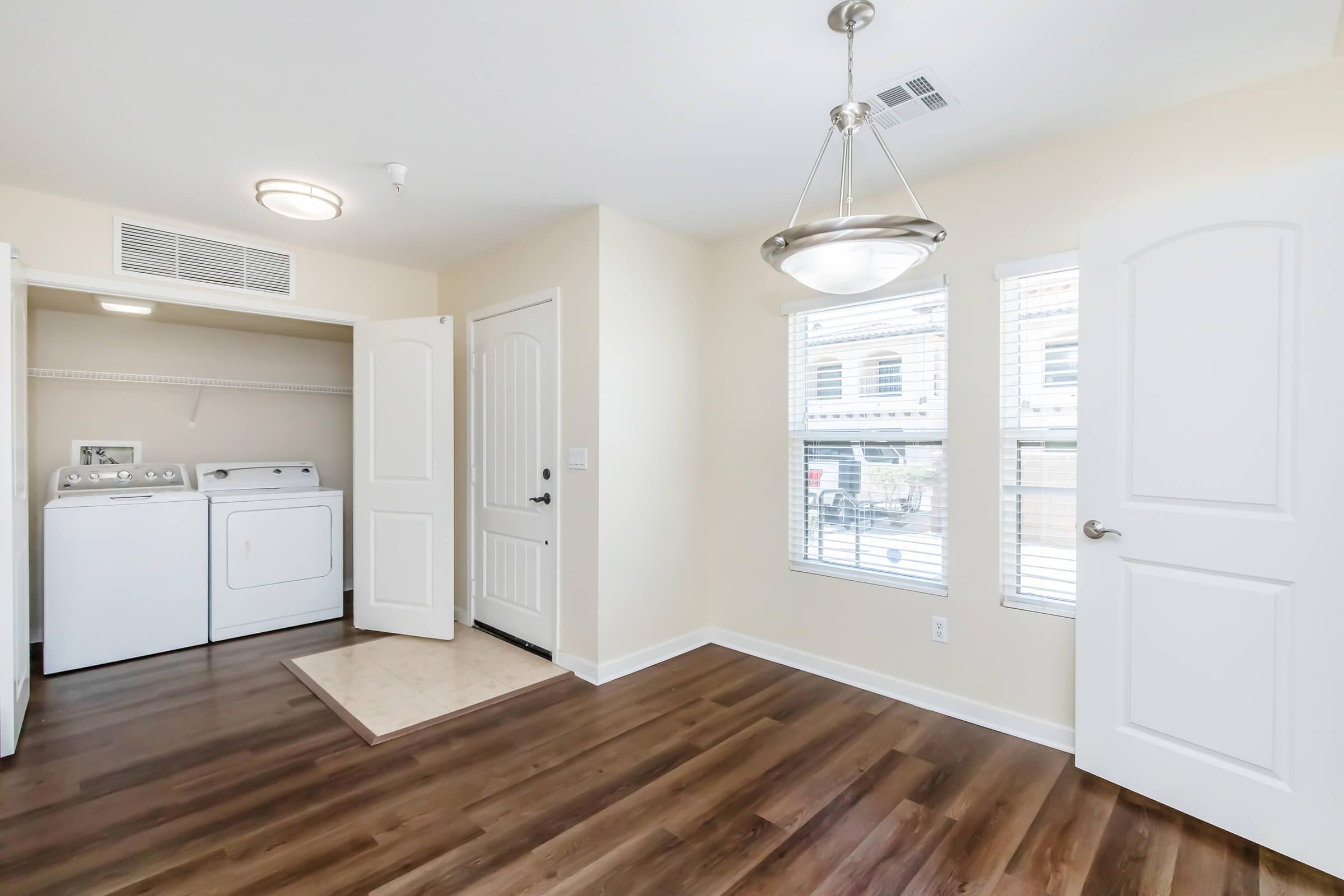A laundry area with a washing machine and dryer, featuring beige walls and hardwood flooring. There are two windows providing natural light, and a chandelier hanging from the ceiling. A door leads to a closet space, enhancing the room's functionality.