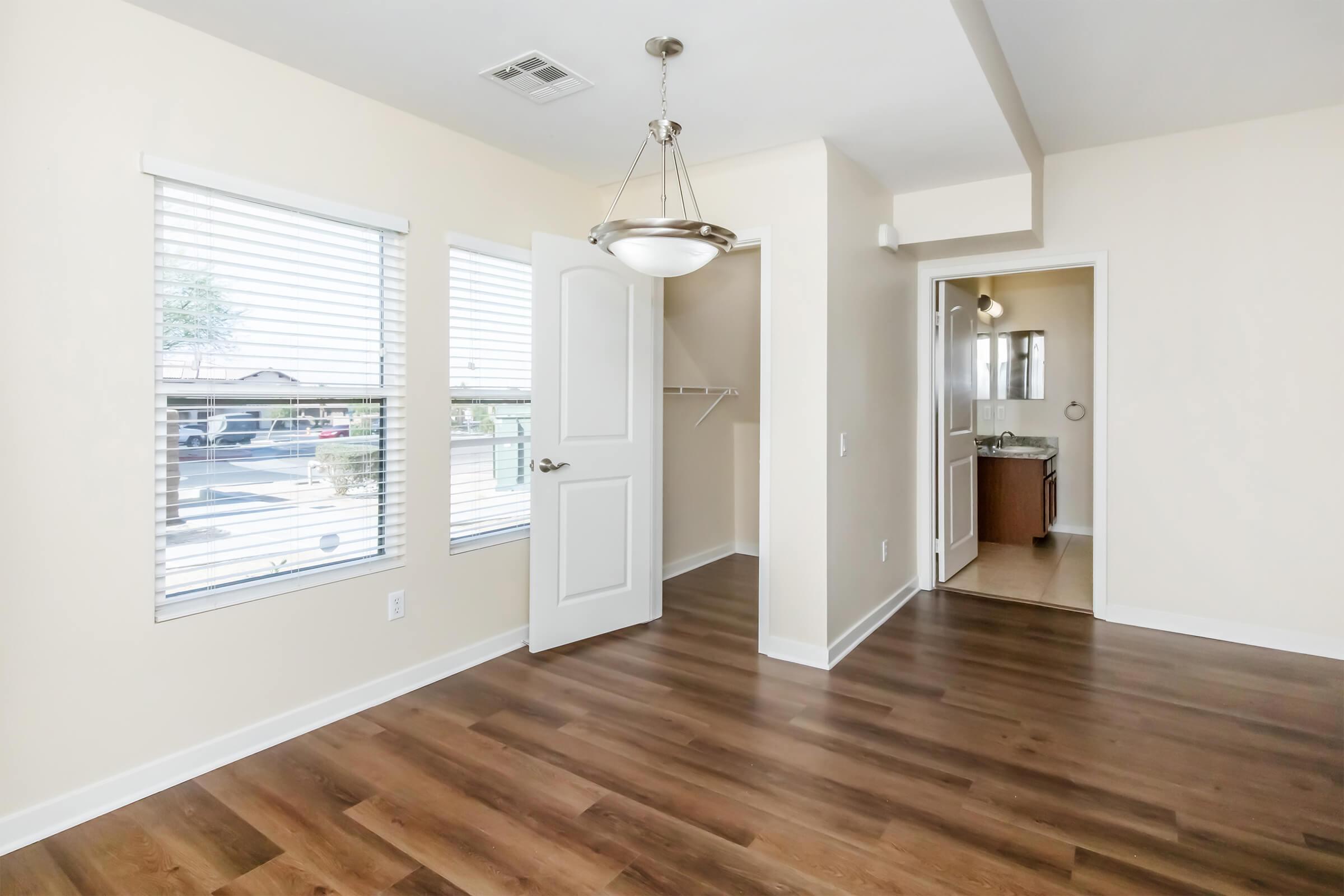 A bright and spacious room with light beige walls and hardwood flooring. It features large windows with white blinds, a ceiling light fixture, and an open doorway leading to a bathroom. A closet space with shelving is visible, creating a cozy atmosphere.