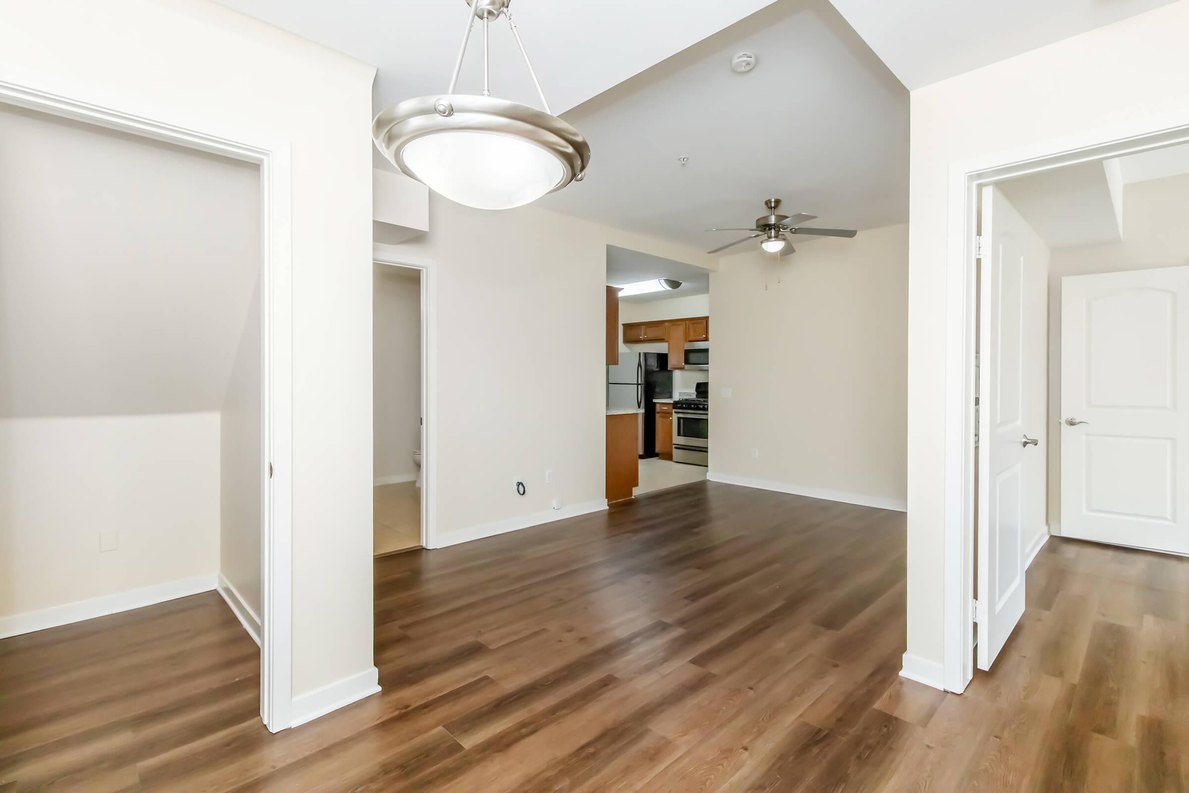 Interior view of an apartment with open layout. Features include a living area with hardwood flooring, ceiling fan, and light fixture. A doorway leads to the kitchen, visible in the background, which has wooden cabinets and appliances. Bright walls and doors create a welcoming atmosphere.