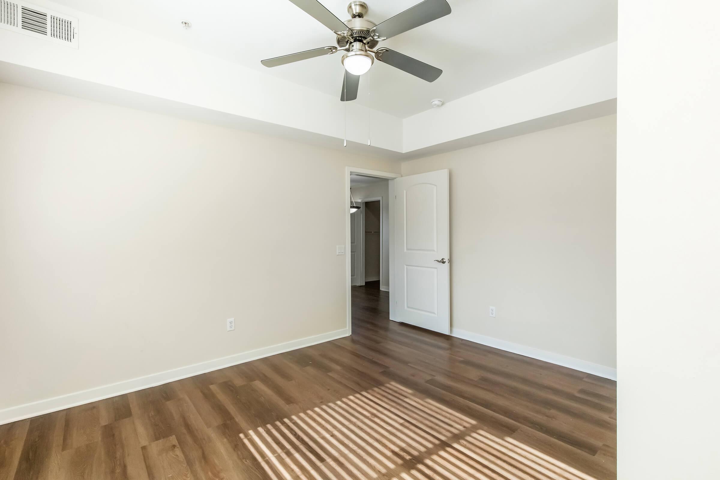 Bright and empty room featuring light-colored walls, a ceiling fan with five blades, and smooth wooden flooring. A door leads to an adjoining space, and sunlight streams through a window, casting striped shadows on the floor. The room has a minimalist design with no furniture.