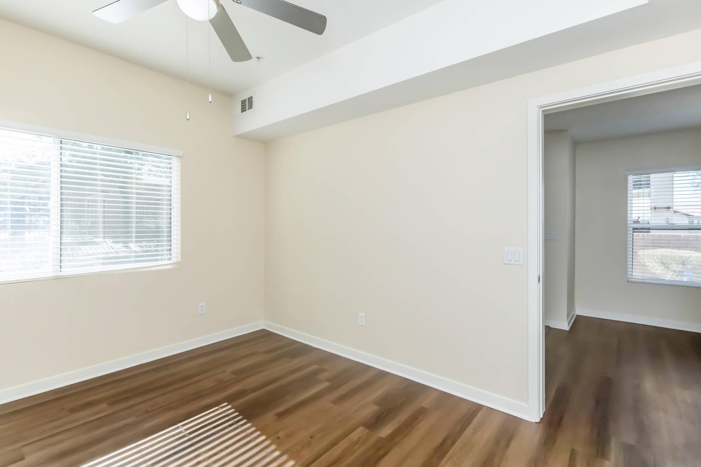 A well-lit interior room featuring light beige walls, a ceiling fan, and a large window with blinds. The floor is made of hardwood, and there is an open doorway leading to another room. Natural light casts a shadow on the floor from the window.