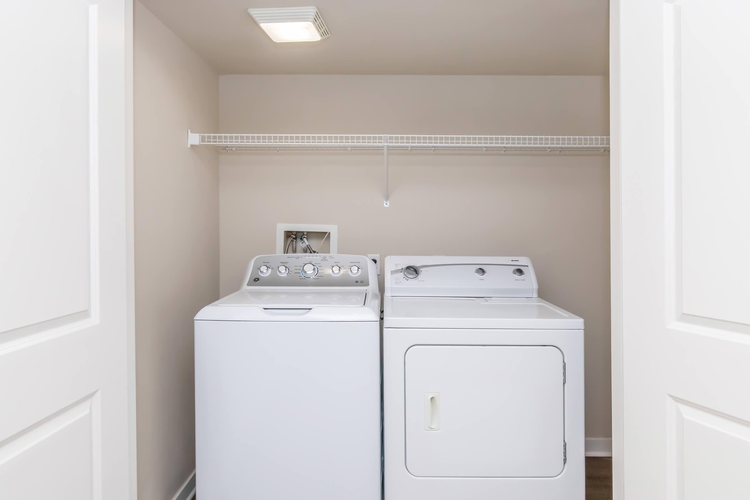 A clean laundry room featuring a washer and dryer side by side, with a shelf above for storage. The walls are painted in a neutral color and there is a light fixture in the ceiling, providing ample lighting. The room is tidy and organized, ideal for laundry tasks.