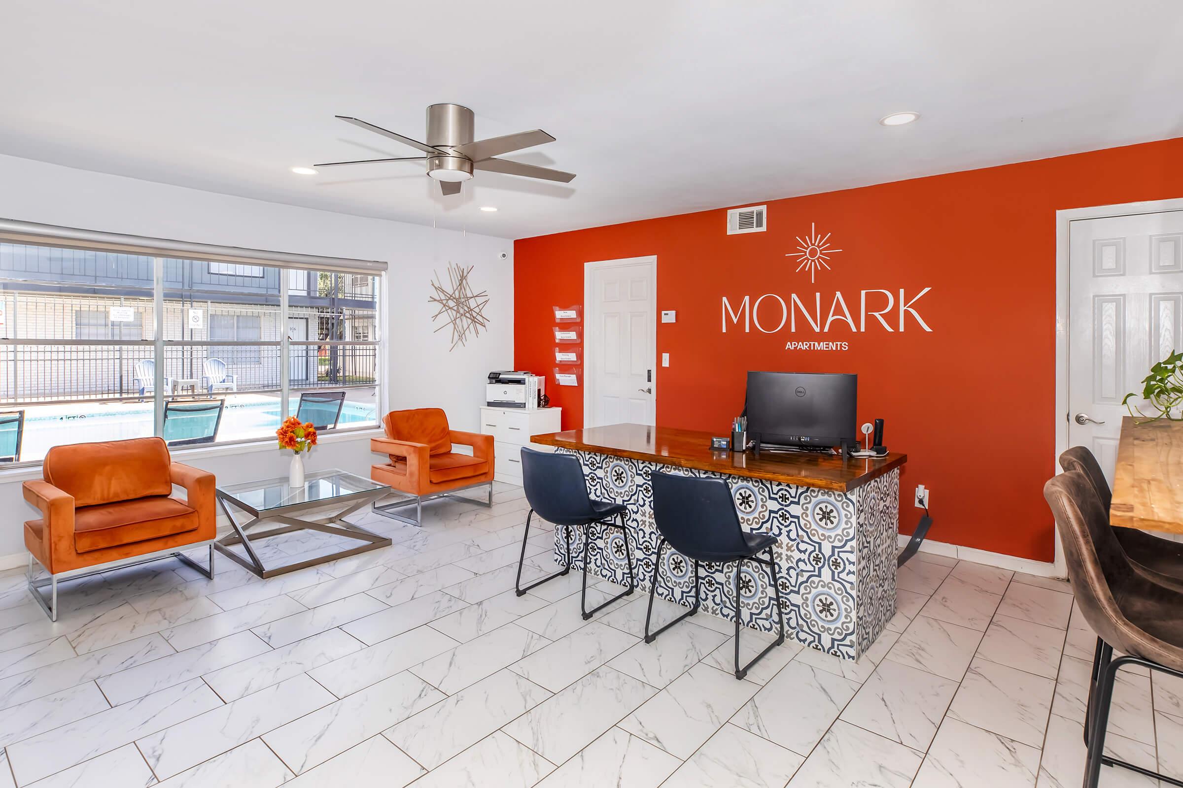 A welcoming reception area featuring a modern desk with a computer, stylish chairs in orange and black, and decorative tile accents. The wall is painted bright orange with the "MONARK APARTMENTS" logo prominently displayed. Large windows provide natural light, and a swimming pool can be seen outside.