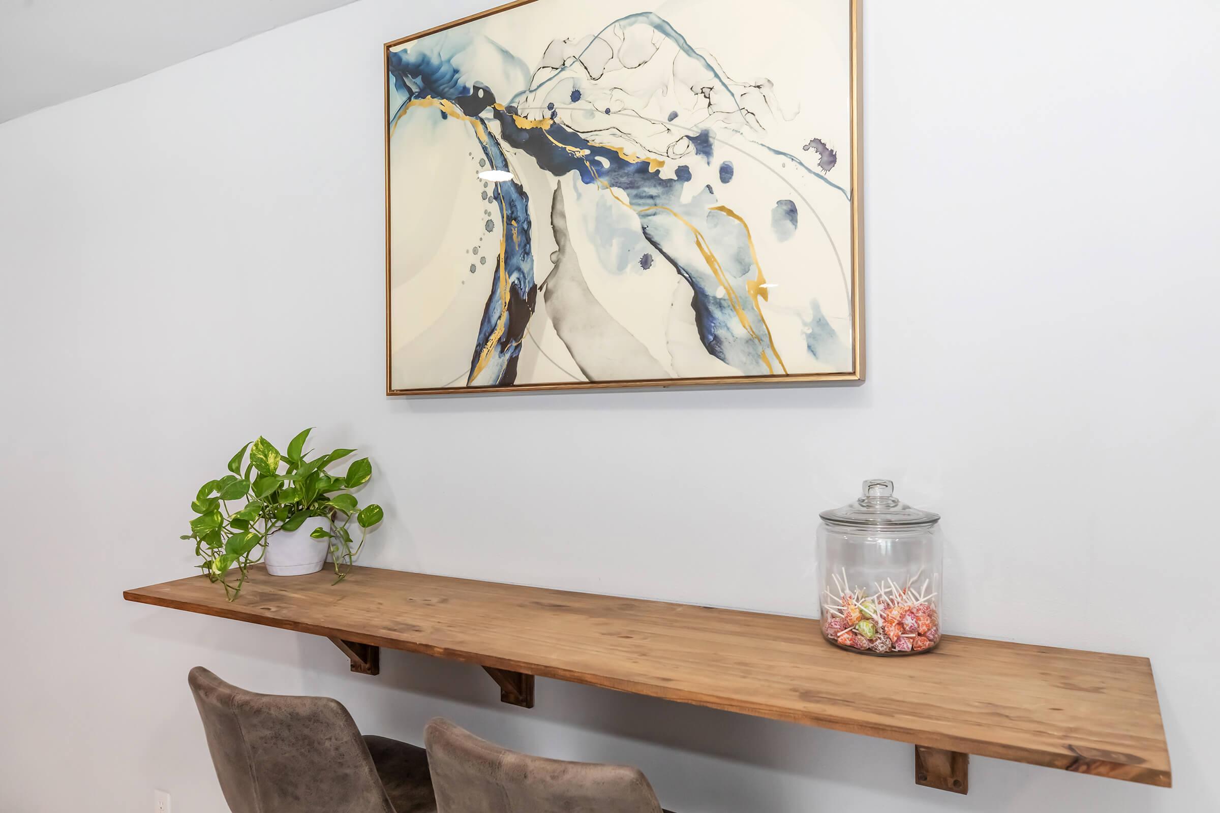 A wooden shelf with two chairs beneath it, adorned with a potted plant and a glass jar filled with colorful candies. Above the shelf, there is an abstract painting featuring blue and gold hues on a light background. The setting has a modern and inviting atmosphere.