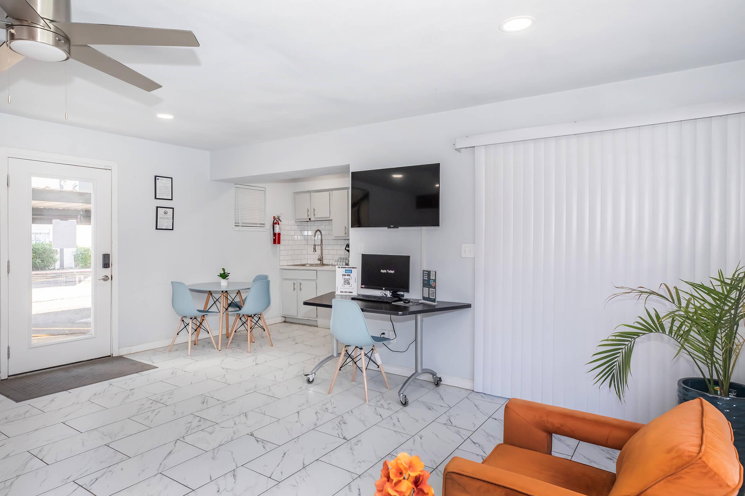 A modern interior space featuring a living area with an orange armchair, a dining area with a small table and blue chairs, and a kitchenette with a television on the wall. Bright, natural lighting highlights the white walls and marble tile flooring. A potted plant adds a touch of greenery to the room.