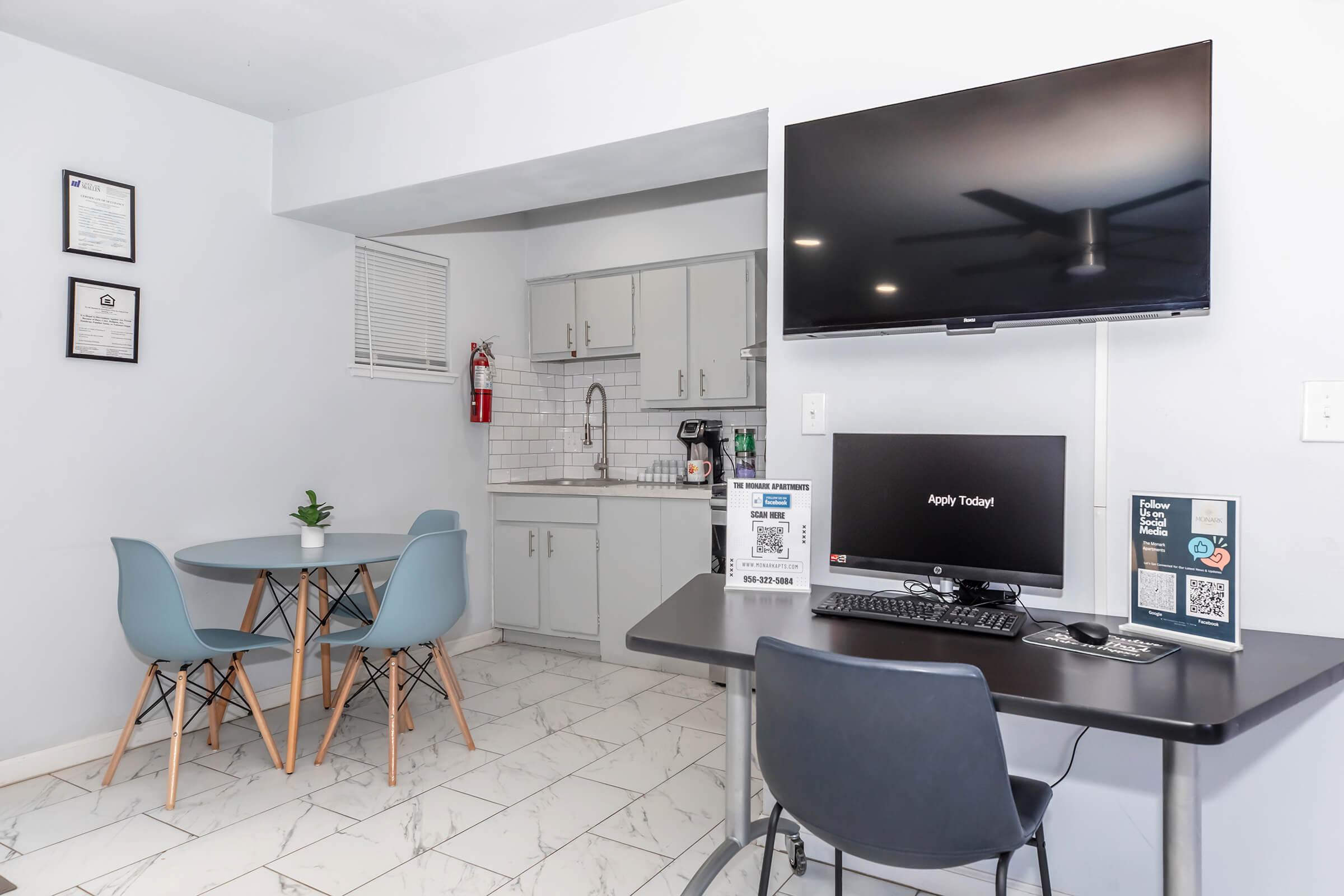 A modern room featuring a small kitchen area with gray cabinets, a sink, and a microwave. There's a table with four chairs, a wall-mounted TV, and a desk with a computer setup. The floor is tiled, and a plant adds a touch of greenery to the space.