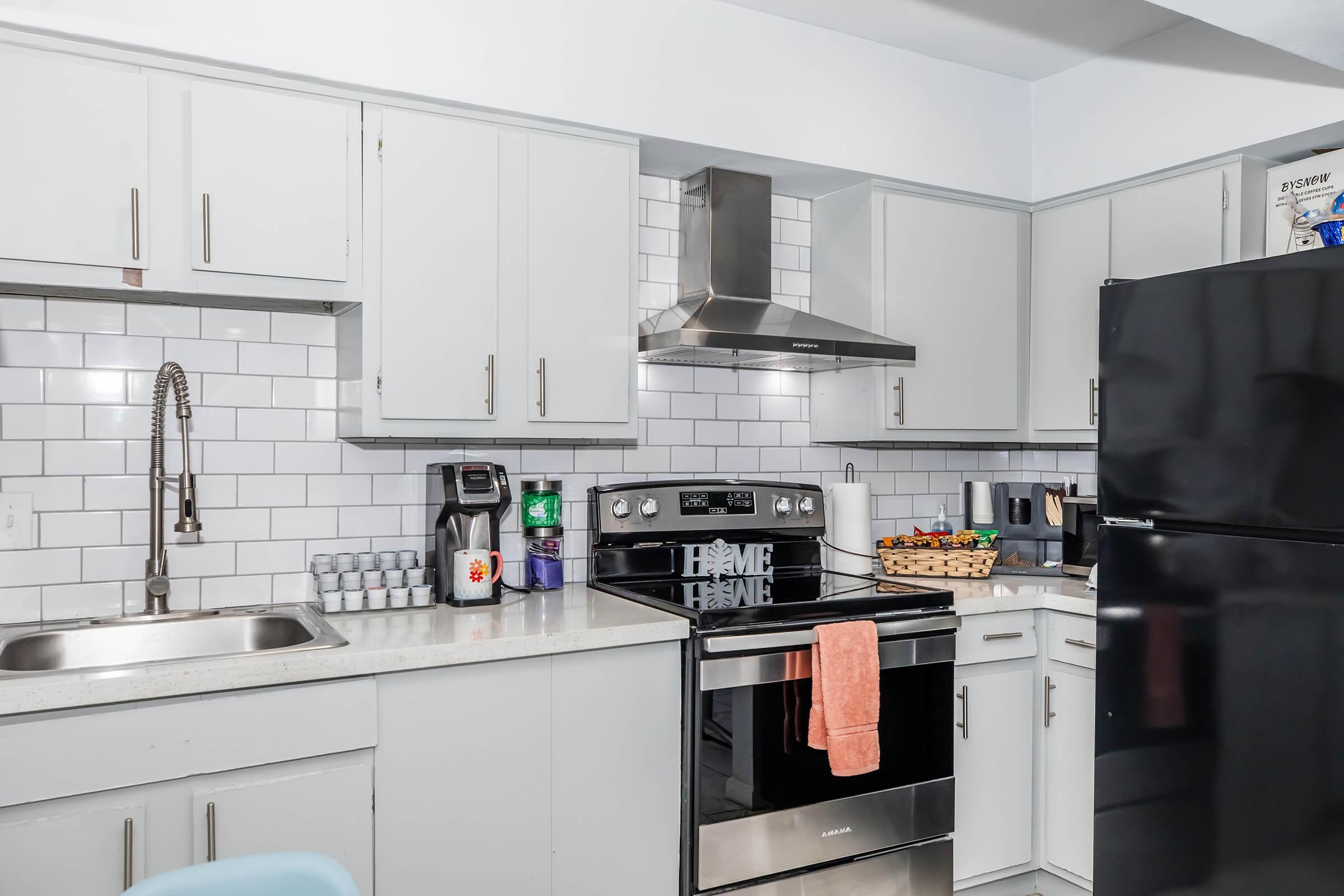 Modern kitchen with light gray cabinets, white subway tile backsplash, an electric stove with an oven beneath, a stainless steel range hood, and a black refrigerator. A silver sink is positioned near the stove, and a coffee maker and a basket of fruit are on the countertop.