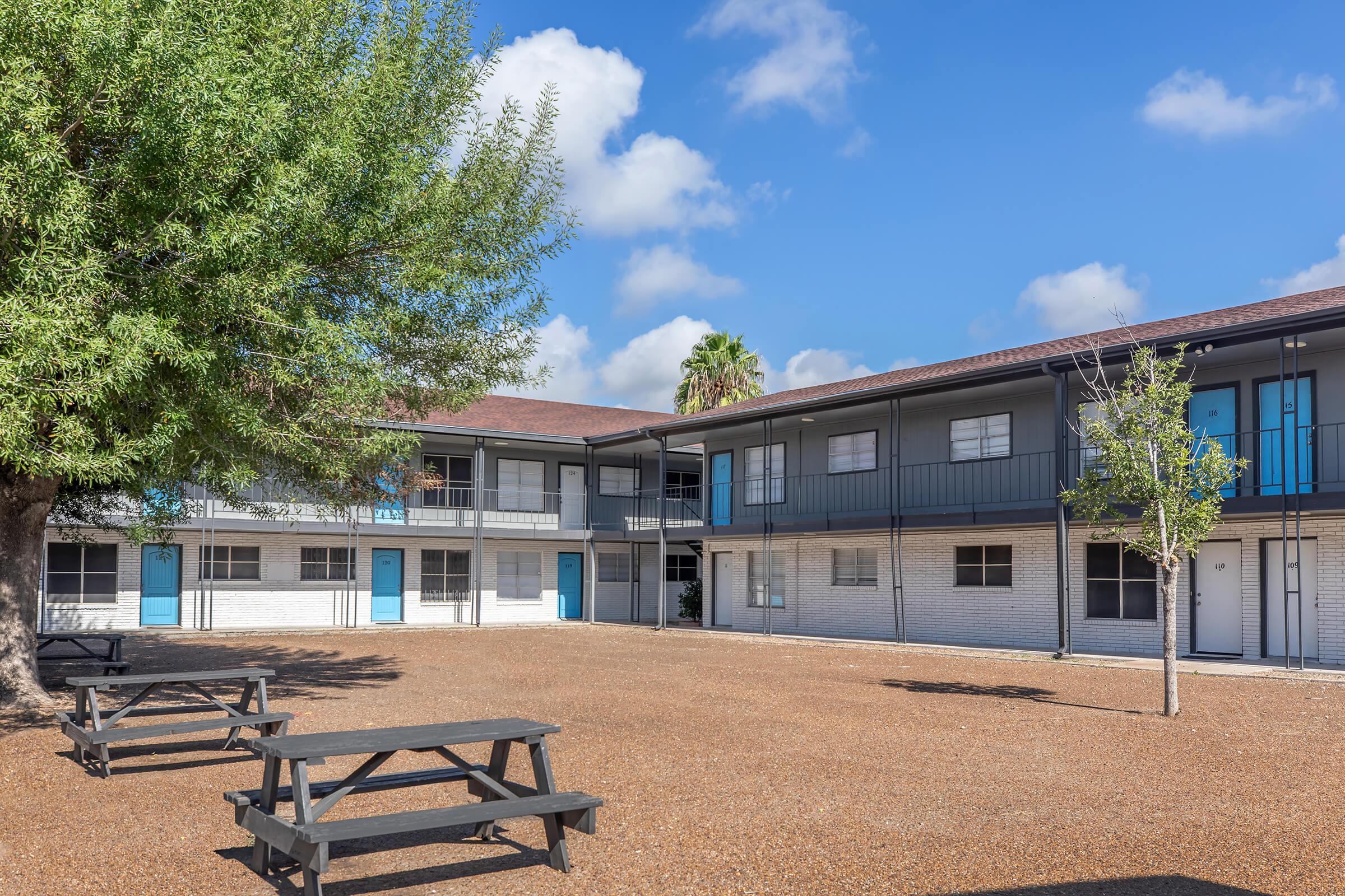 A courtyard view of a multi-story building with blue doors and windows, surrounded by gravel and a few trees. Two picnic tables are placed in the foreground, providing a recreational area. The sky is clear with scattered clouds, creating a bright, inviting atmosphere.