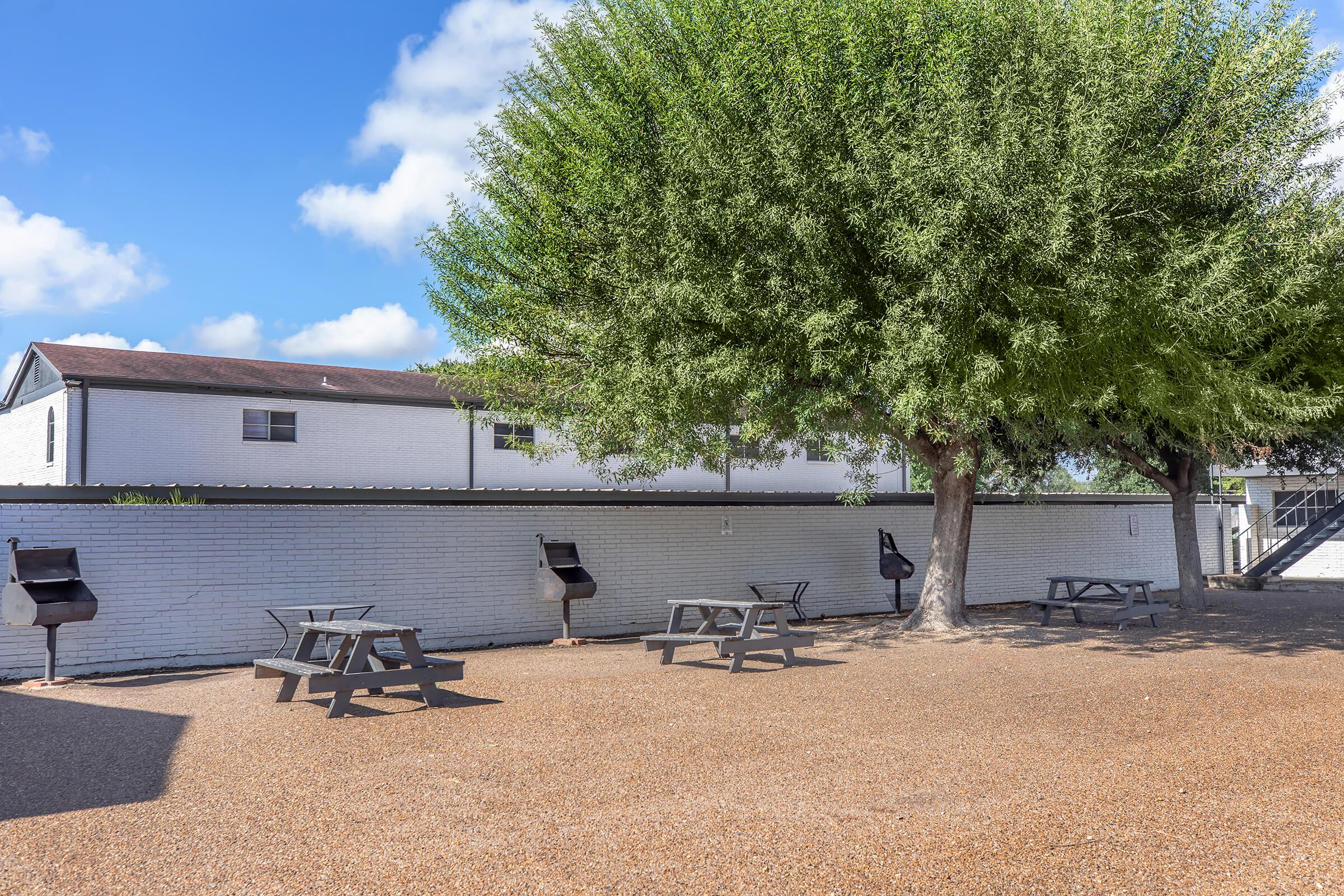 Outdoor area featuring several picnic tables and two grill stations under the shade of a large tree. A white brick wall borders the space, with a building visible in the background. The ground is covered in gravel, and the sky is partly cloudy, creating a pleasant environment for gatherings or relaxation.