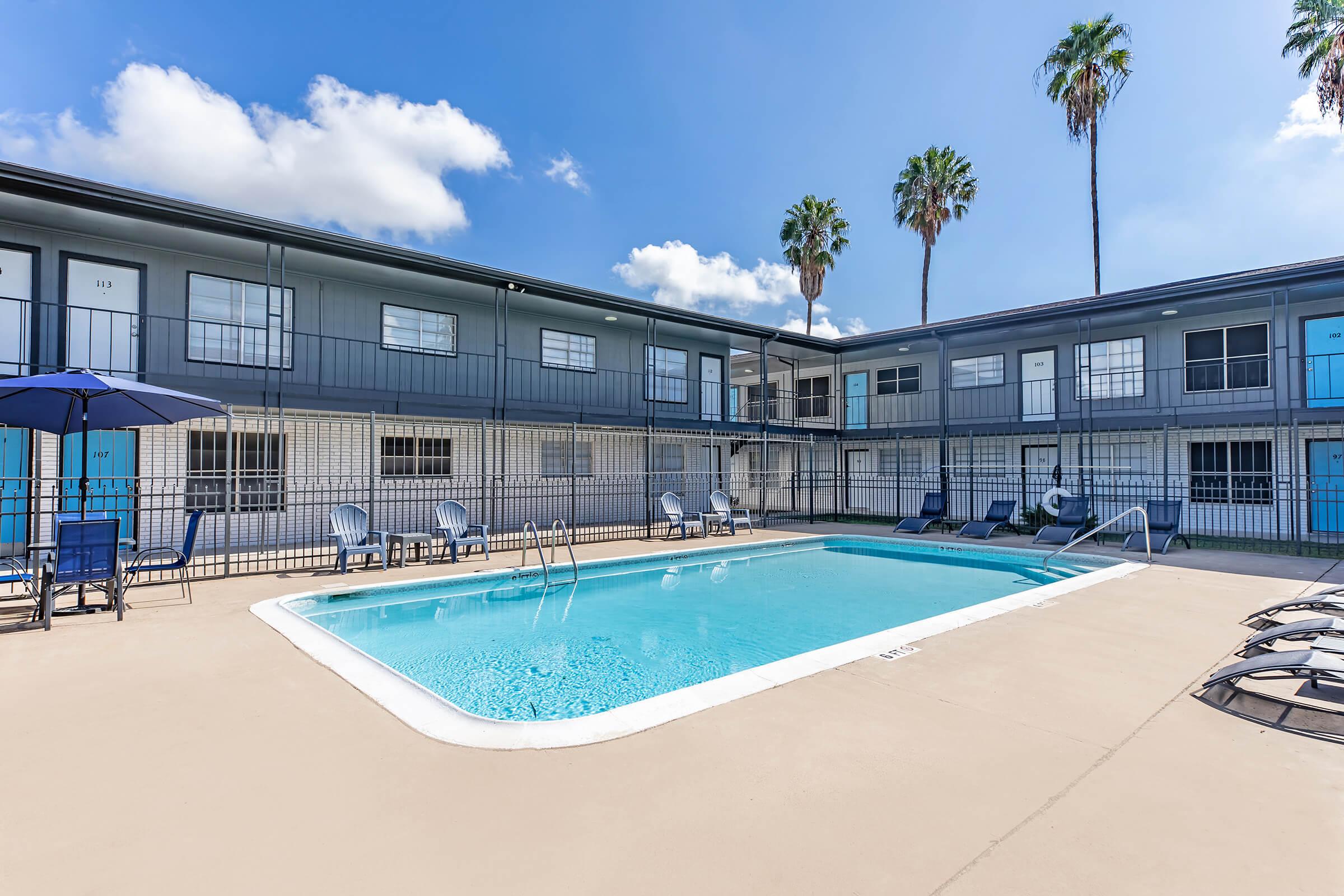 A clear swimming pool surrounded by lounge chairs, with a few palm trees in the background. The pool area is enclosed by a fence, and the adjacent building features multiple balconies with blue doors. The sky is bright with some clouds, creating a relaxed atmosphere.