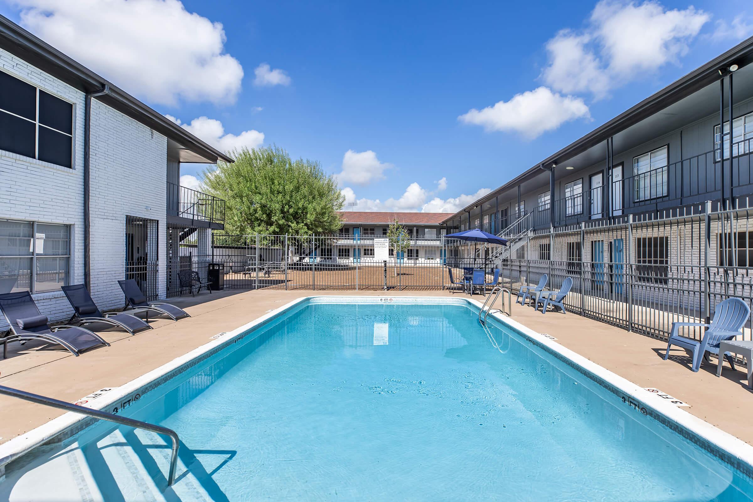 A clear swimming pool with a ladder, surrounded by lounge chairs, set in a courtyard between two multi-story apartment buildings. The sky is blue with a few clouds, and there is a tree providing shade in the background.