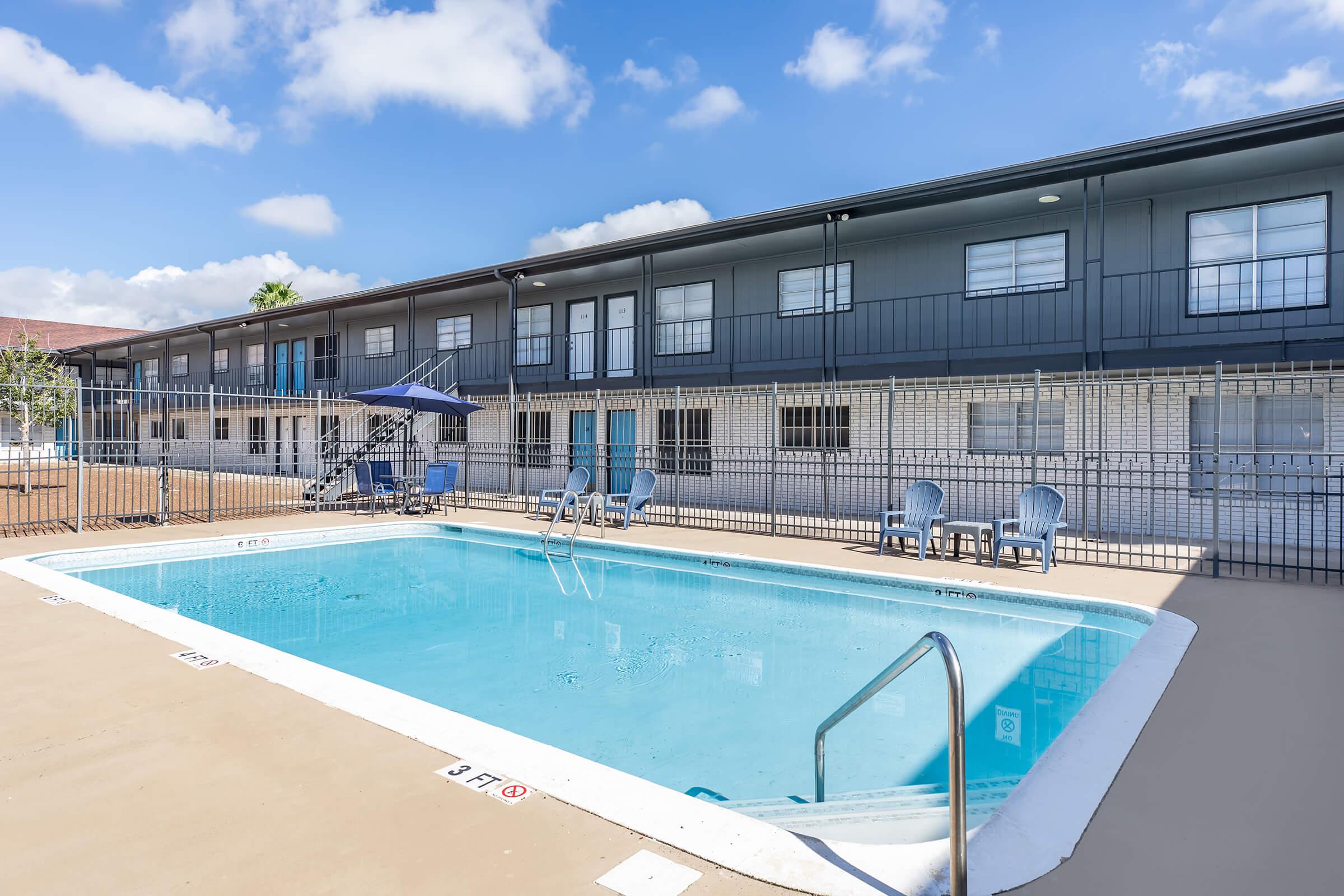 A clean outdoor swimming pool surrounded by a fence, with lounge chairs and umbrellas nearby. In the background, there are modern apartment buildings with balconies and a clear blue sky overhead.