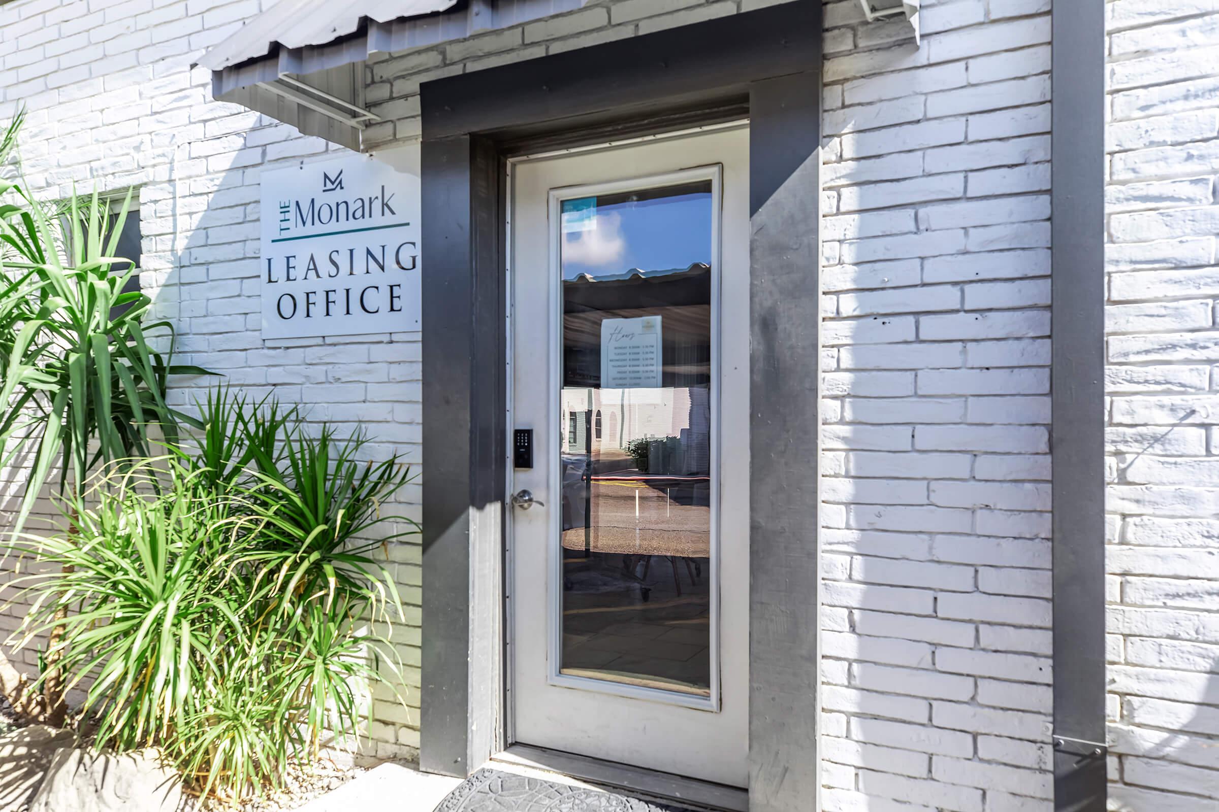 Exterior view of the leasing office for The Monark, featuring a glass door framed in black, with a white brick wall and a sign that reads "Leasing Office." Lush green plants are visible near the entrance, and a shaded area can be seen through the door.