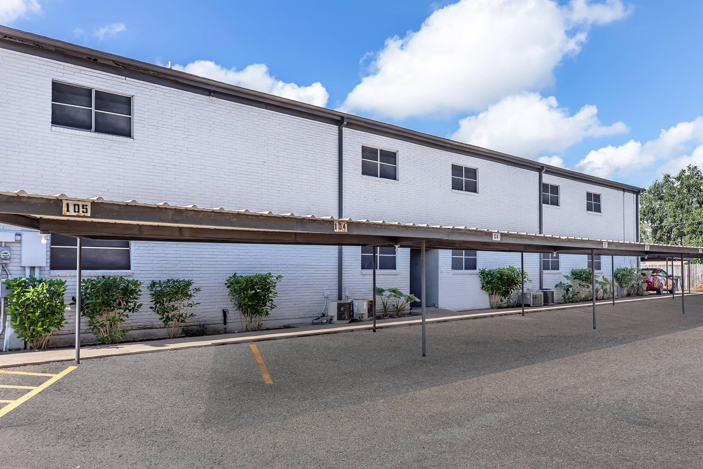 A row of covered parking spaces in front of a two-story white building with multiple windows. The parking area is neatly organized, with numbered spaces visible. Lush green bushes line the building, and the sky is clear with a few clouds.