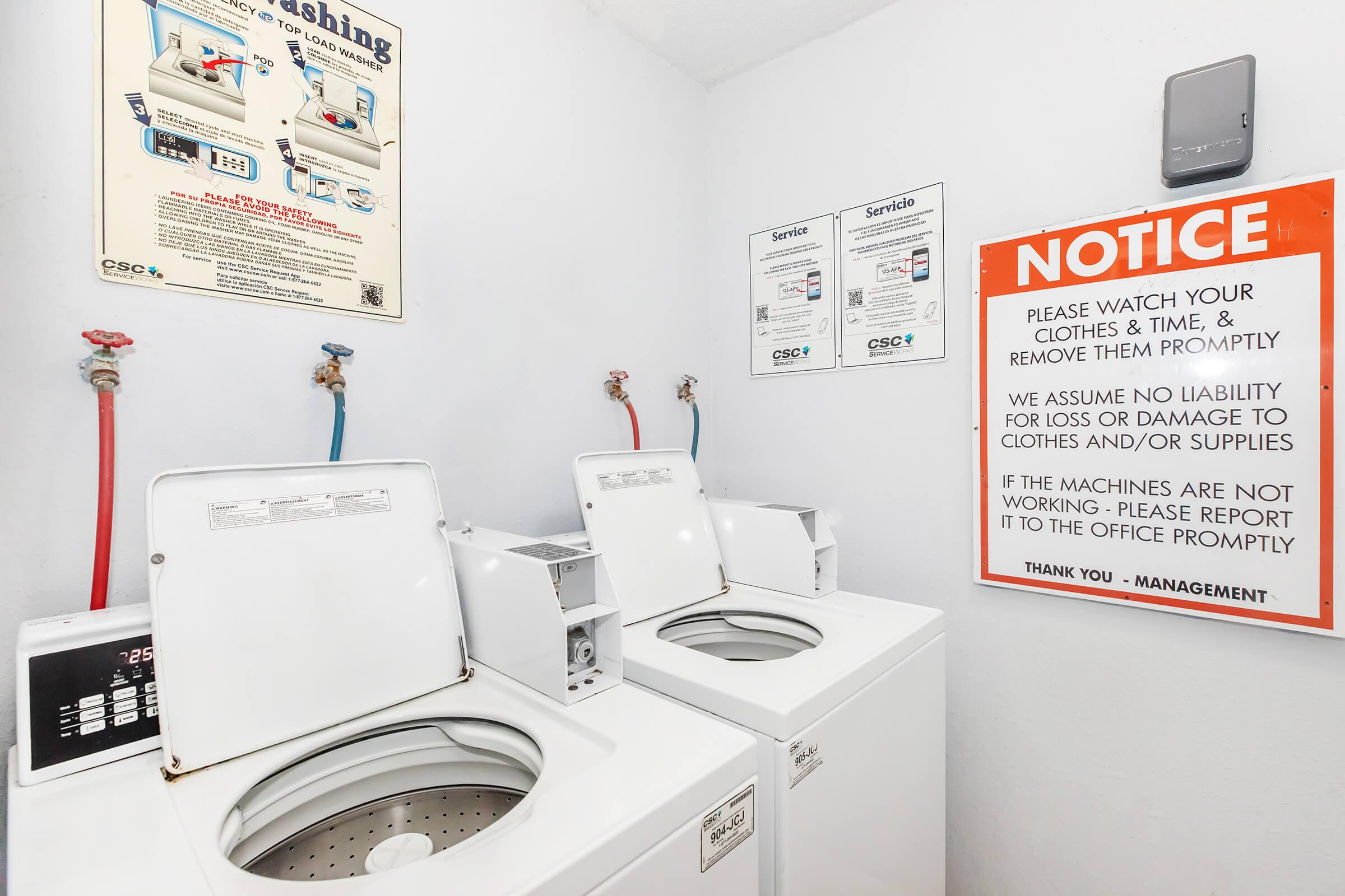 Two white washing machines are positioned side by side in a laundry room. Above them, a notice warns users to watch their clothes and report any machine issues promptly. Informational posters about laundry services are on the wall. The overall environment is clean and functional.