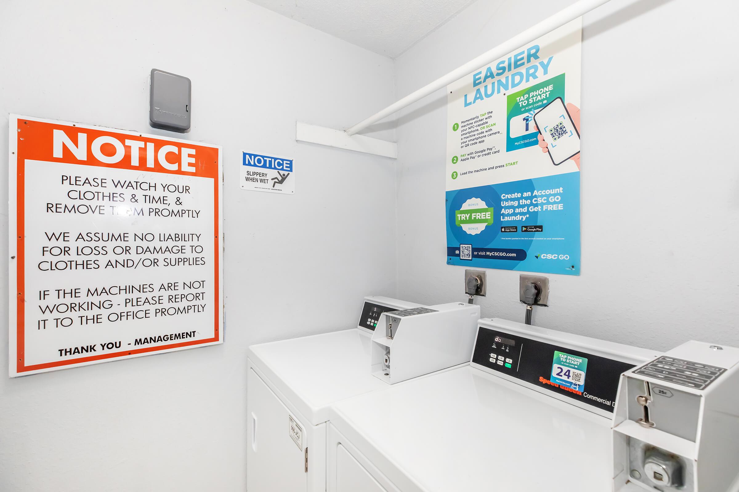A laundry room featuring two coin-operated washing machines and a notice board. The sign emphasizes the importance of watching clothes and timely removal, and includes a notice about liability for loss or damage. A bright poster advertises an easier laundry service option with instructions for use.