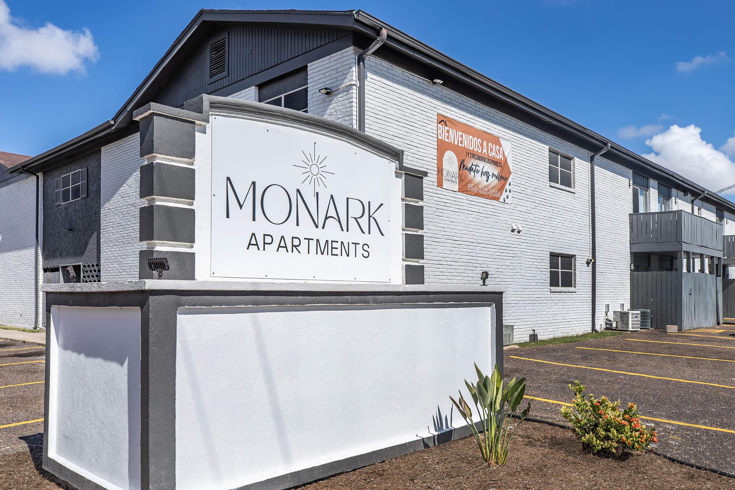 Sign for Monark Apartments, featuring the name prominently. The building has a modern design with a light-colored exterior and blue sky in the background. There is a banner on the upper right indicating an announcement or event. Landscaping includes low shrubs in front of the sign.