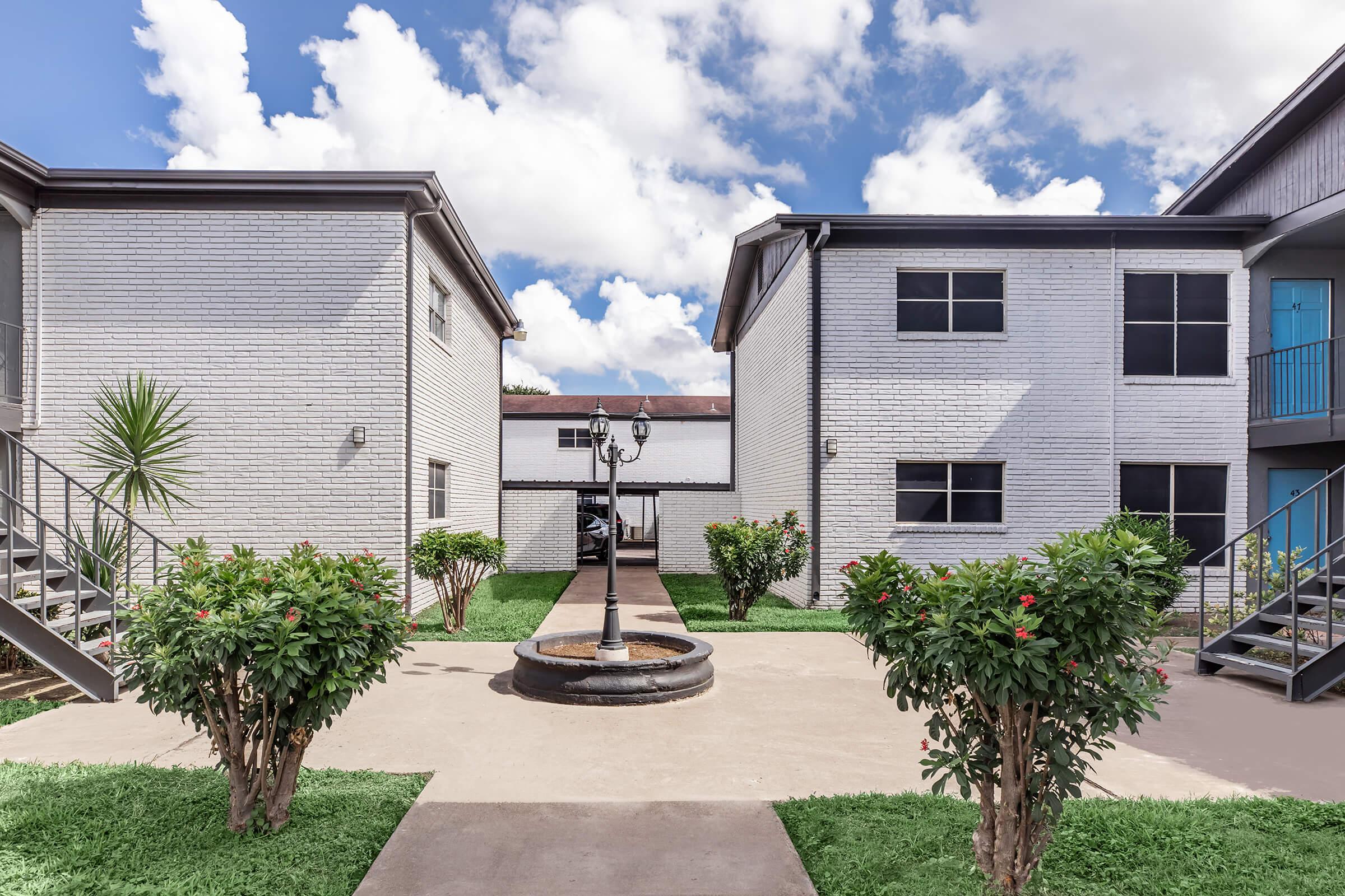 A well-maintained courtyard featuring two white brick apartment buildings. The area is landscaped with green shrubs and grass, with a central circular fountain and a pathway leading between the buildings, under a partly cloudy sky.