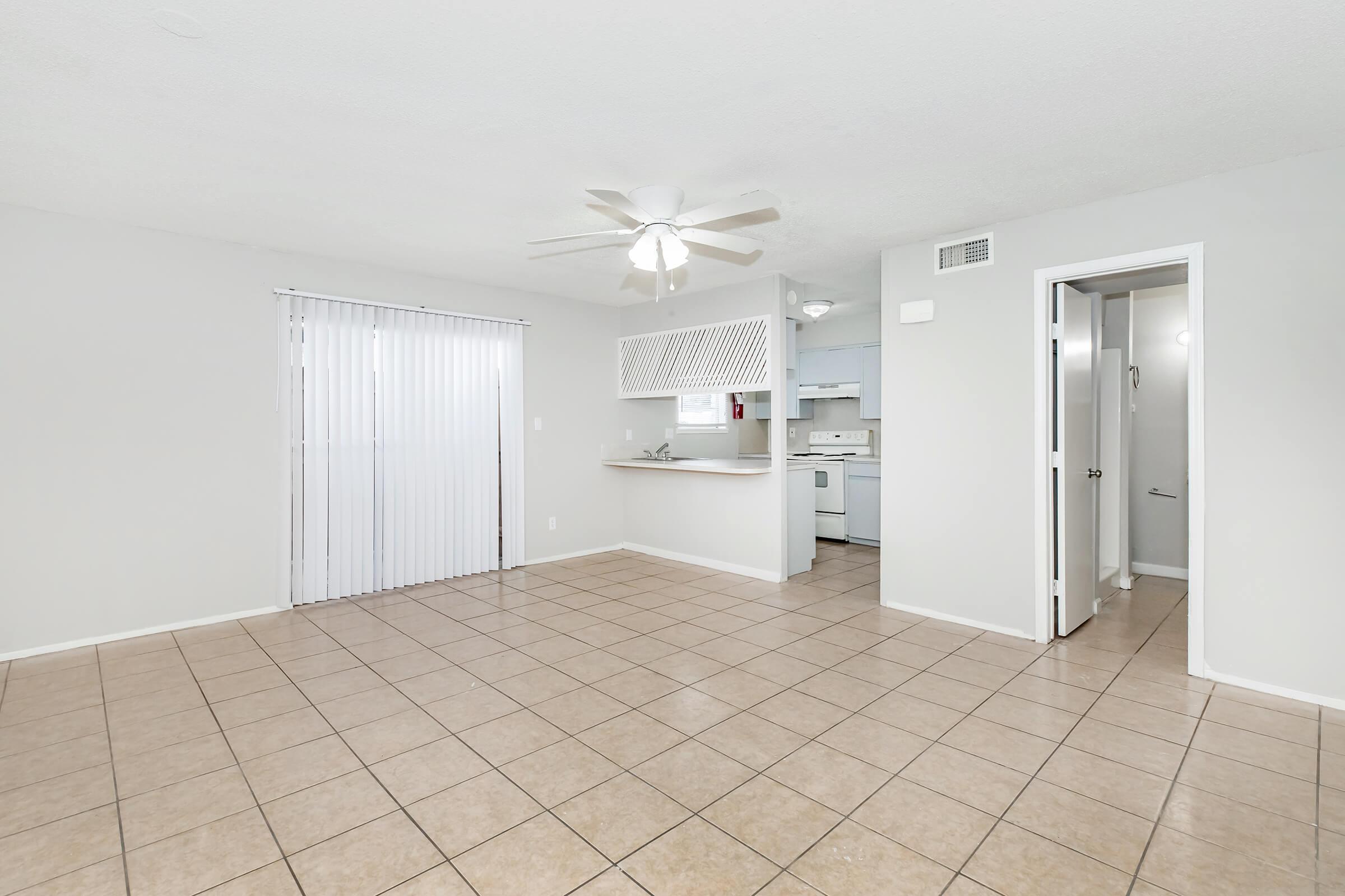 Open concept living area with tiled flooring, ceiling fan, and sliding glass door leading to an outdoor space. In the background, a kitchen with white appliances is visible. A doorway on the right leads to another room. The walls are painted light gray, creating a bright and spacious atmosphere.