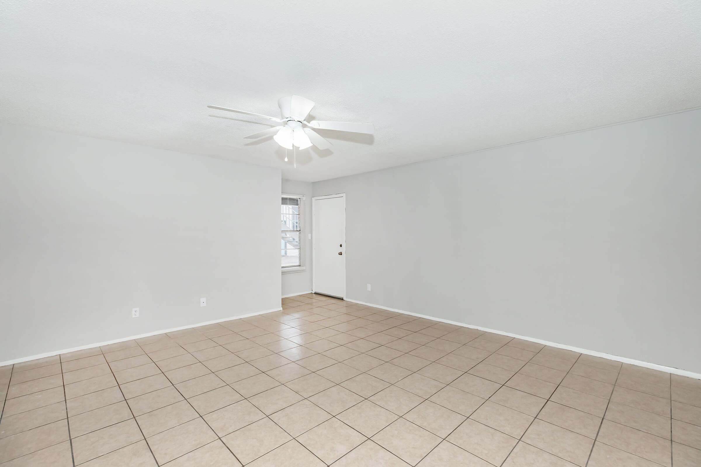 A spacious, empty room with light gray walls and beige tiled flooring. A ceiling fan is mounted on the ceiling, and there's a door leading outside. The area is well-lit with natural light from a window, creating an open and airy atmosphere.