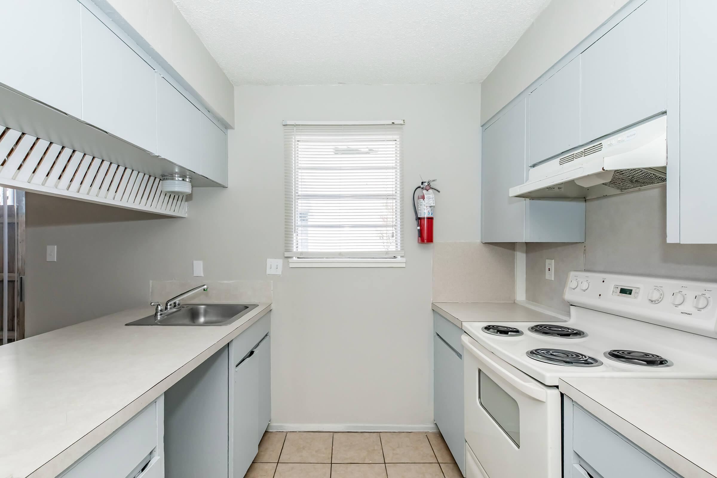 Bright, modern kitchen featuring light blue cabinets, a stainless steel sink, and white appliances, including an electric stove and oven. A window provides natural light, and a fire extinguisher is mounted on the wall for safety. The tile floor complements the clean, contemporary design.
