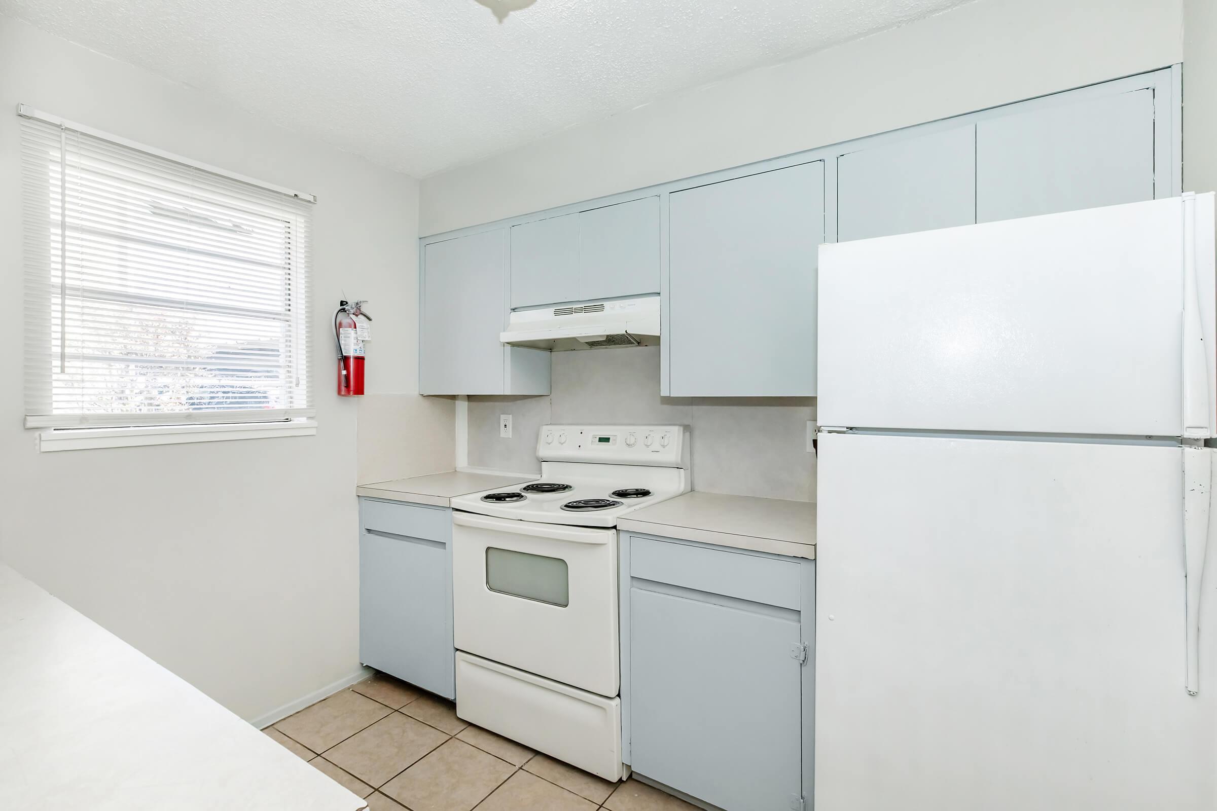 A clean, simple kitchen featuring light gray cabinets, a white stove, and a refrigerator. The countertop is empty, and there is a window with blinds on the left side, allowing natural light. A fire extinguisher is mounted on the wall for safety.