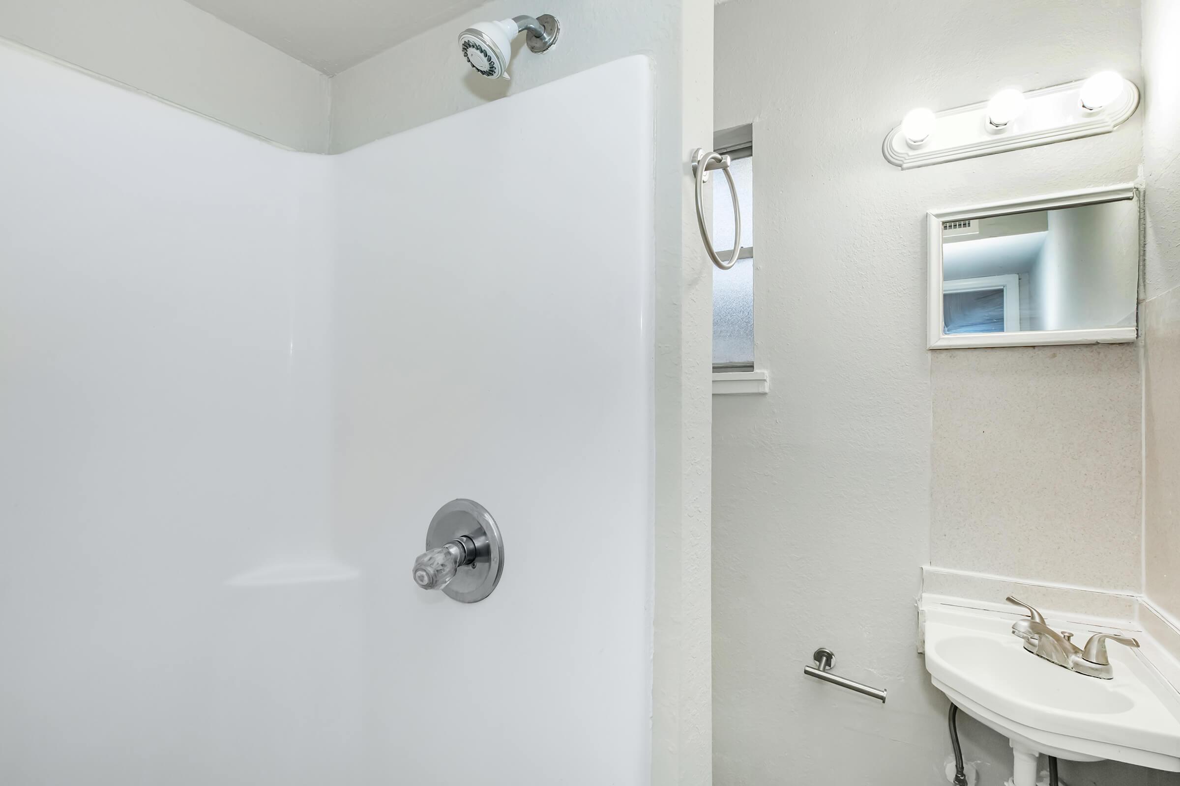 A modern bathroom featuring a white shower stall, a small vanity with a sink and mirror, and neutral-colored walls. There is a towel bar beside the sink and overhead lighting, creating a bright and clean atmosphere.