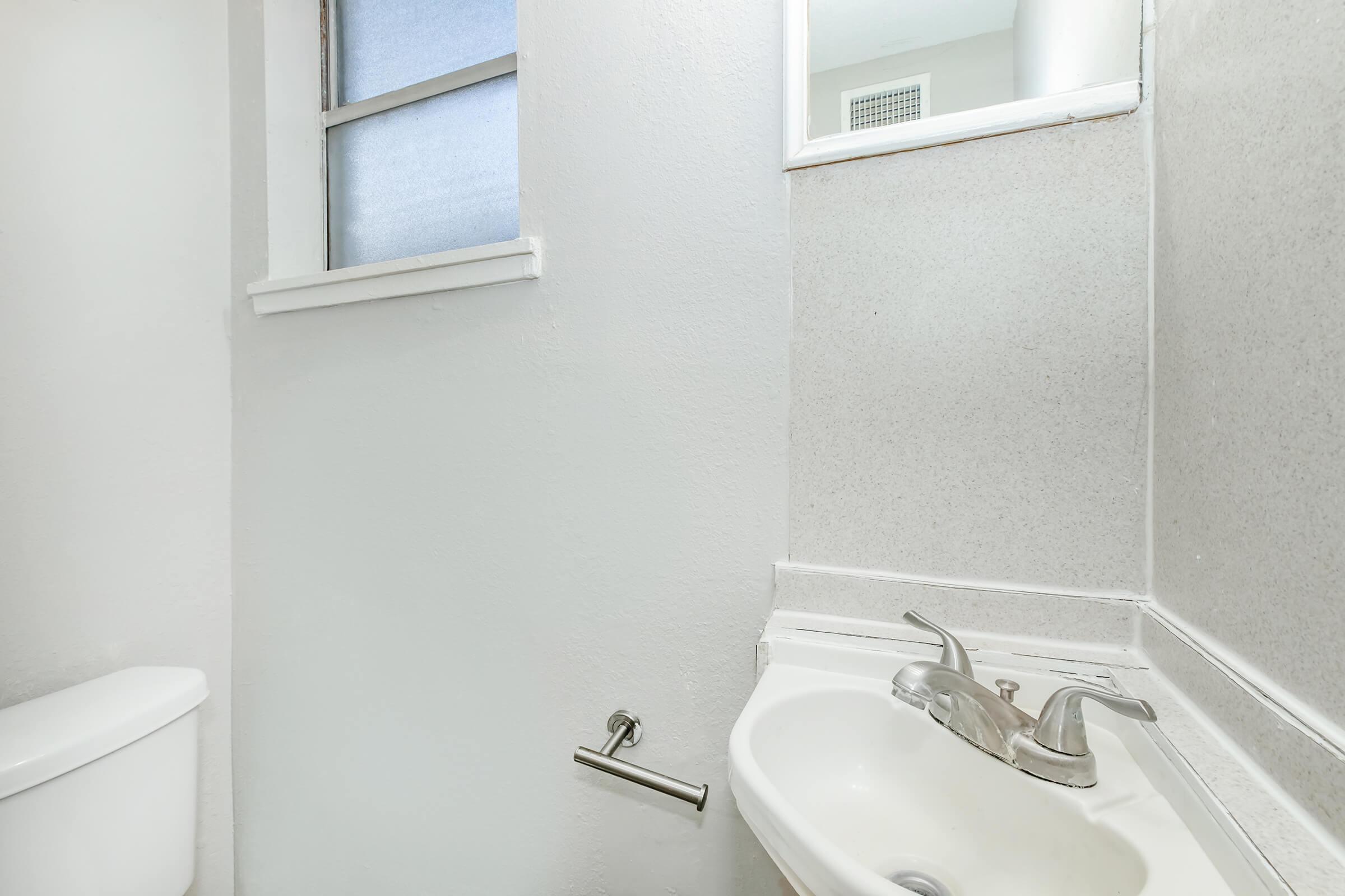A small bathroom featuring a toilet, a sink with a faucet, and a wall-mounted mirror. The walls are painted white, and natural light enters through a small window with a frosted glass panel. The overall atmosphere is simple and functional.