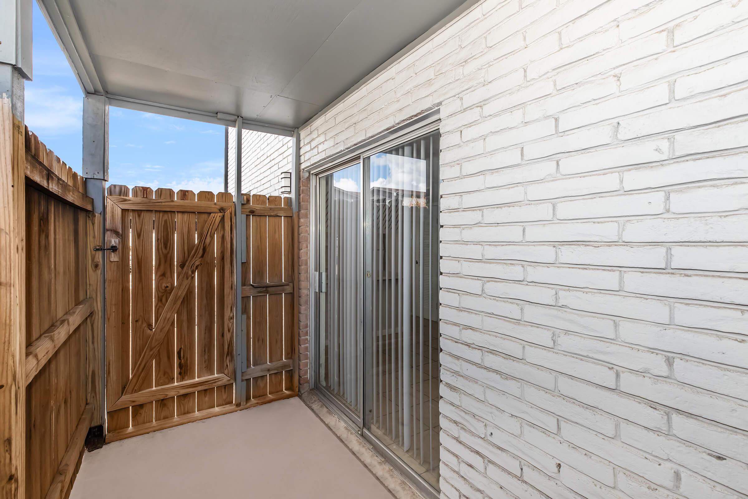 A small outdoor patio area with a brick wall and a wooden fence. There is a glass sliding door leading into an interior space. The flooring is plain and the area is well-lit, with blue sky visible above.