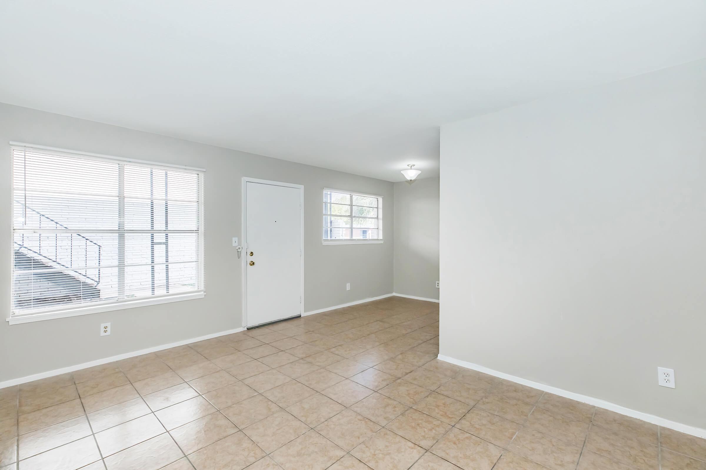 An empty room with light-colored walls and tile flooring. It features a single front door, two windows with blinds allowing natural light, and a ceiling light fixture. The space is minimalistic and uncluttered, suggesting potential for various furniture arrangements.