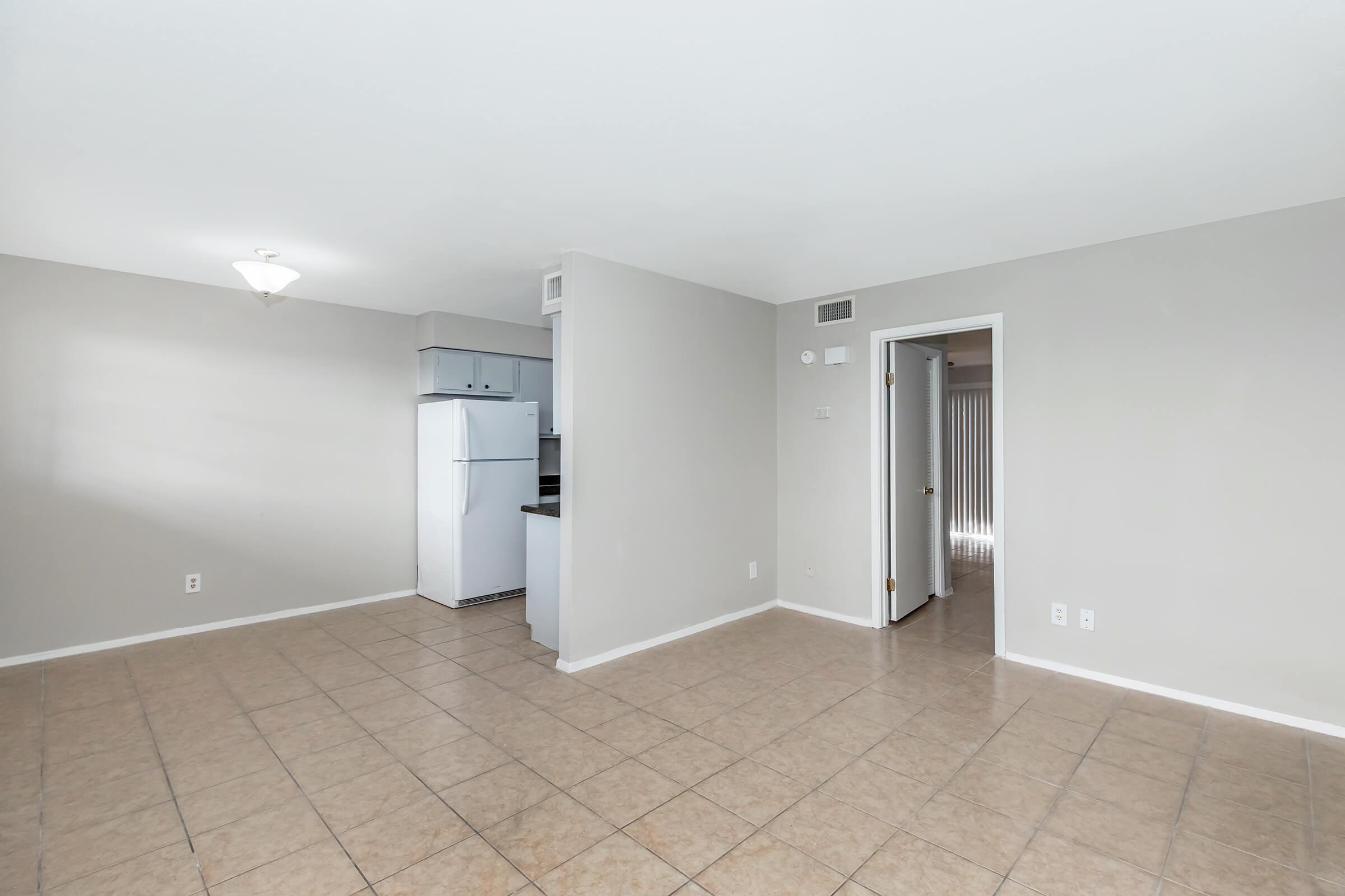 Open-concept living space with beige tiled flooring, light gray walls, and a ceiling light fixture. On the left, a small kitchen area with a white refrigerator and dark countertops is visible. A door on the right leads to another room. The area is well-lit and spacious.