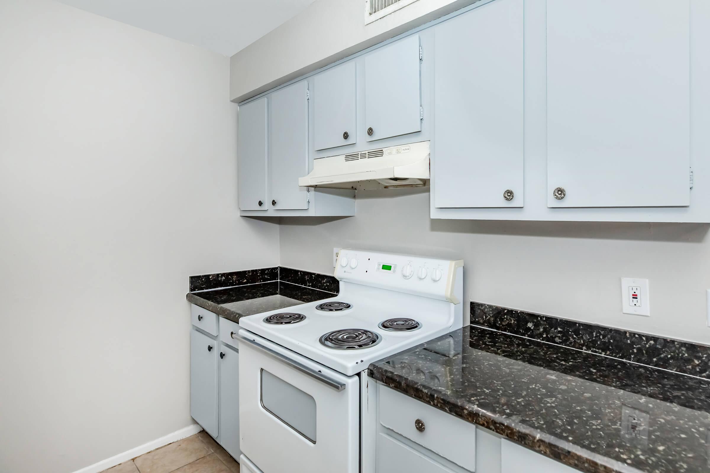 A modern kitchen featuring light blue cabinets, a white stove with four burners, an overhead exhaust hood, and dark granite countertops. The walls are painted a neutral light gray, and the flooring is tiled.