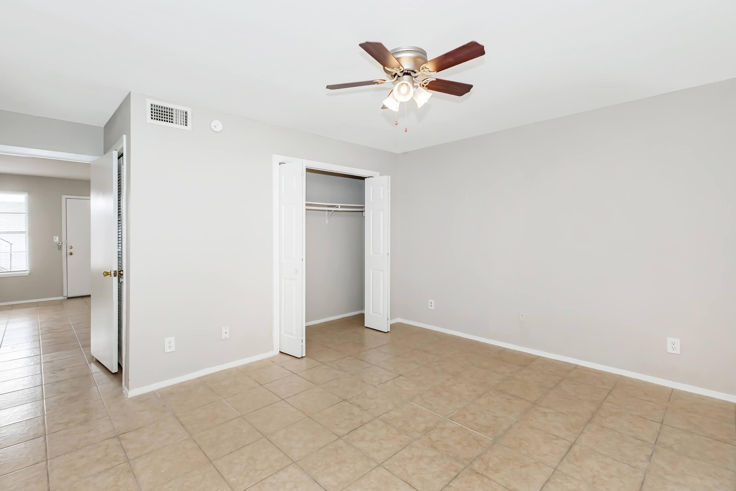 A spacious, empty room with a light-colored wall, tiled flooring, and a ceiling fan. One corner features a closet with double doors, and an entrance leads to another area, suggesting a well-lit, open living space.