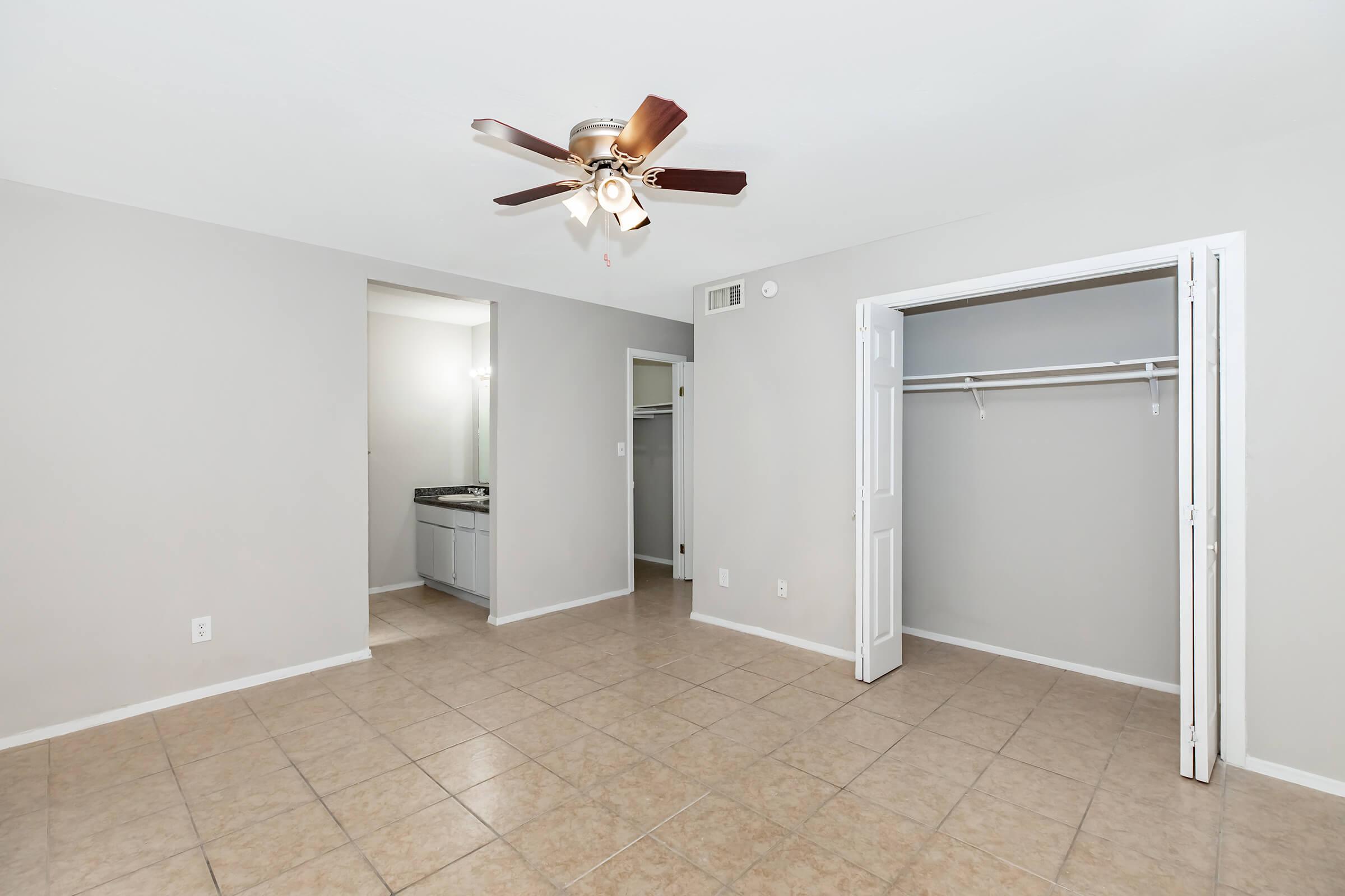 Empty room featuring beige tiled flooring, a ceiling fan with wooden blades, and light gray walls. There are two doorways leading to a bathroom and a closet, with a built-in shelf in the closet. The room is well-lit with natural light.