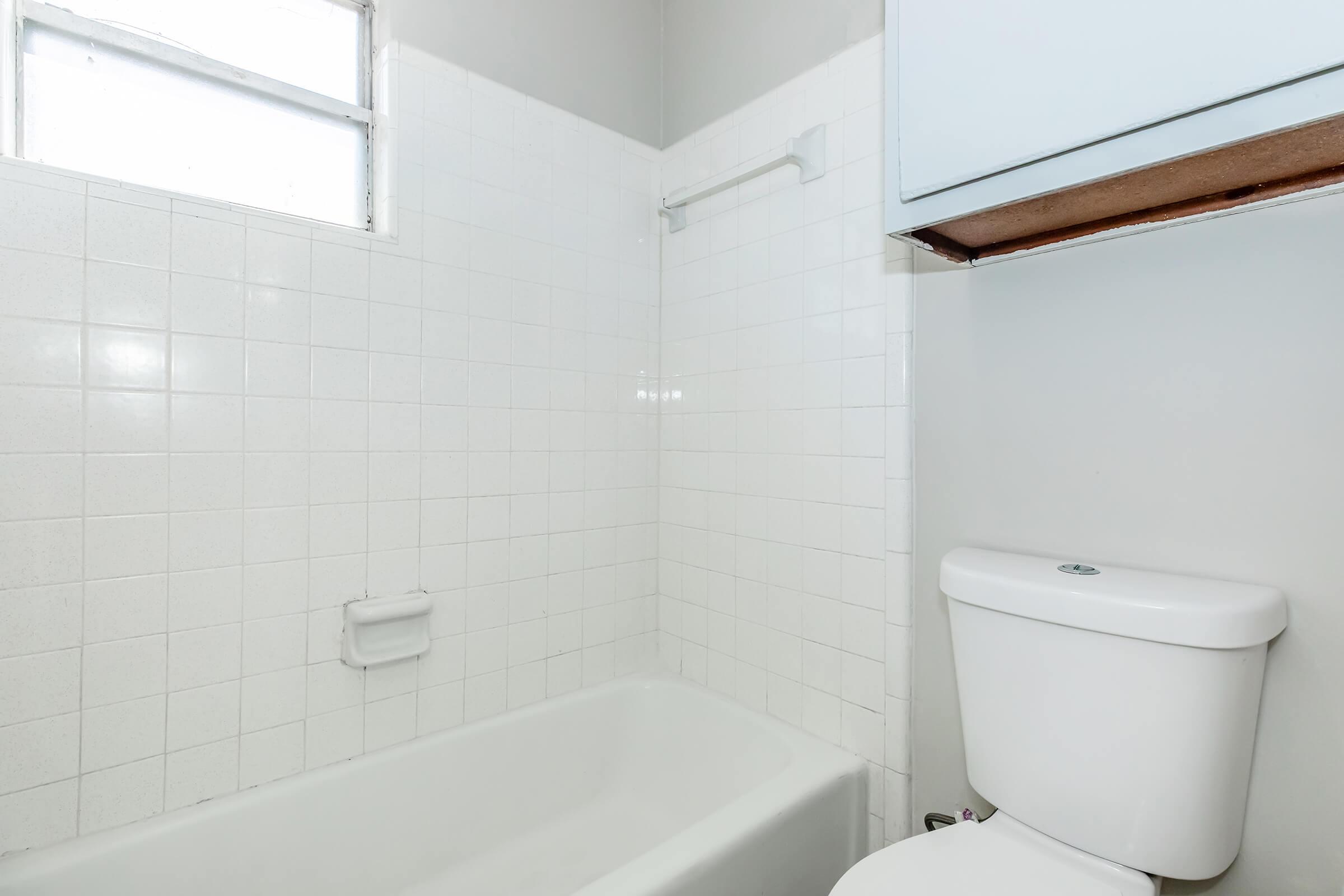 A clean, minimalistic bathroom featuring a white bathtub and a toilet. The walls are tiled with white square tiles, and there is a small window allowing natural light. Above the bathtub, a showerhead is mounted, and a cabinet is visible above the toilet. The overall design is simple and functional.