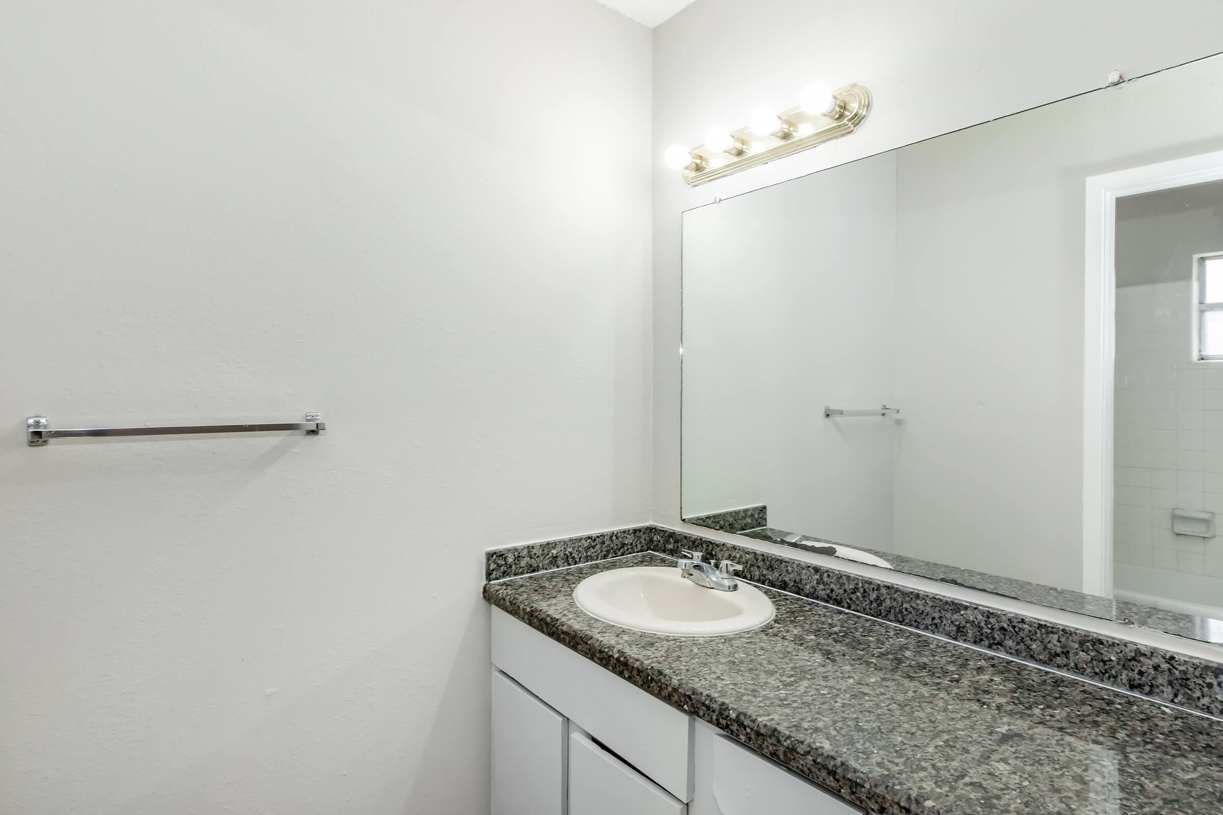 A simple bathroom featuring a light-colored wall, a granite countertop with a sink, and a large mirror above the sink. A towel bar is mounted on the wall, and there's a doorway visible leading to another room. The overall setting is bright and clean.