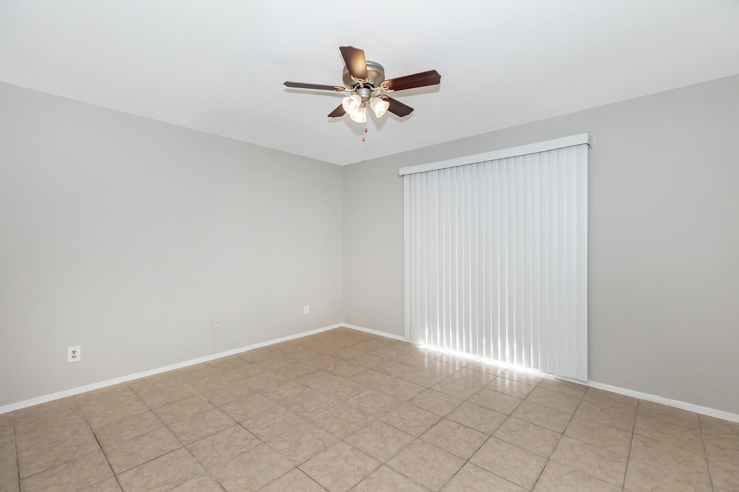 A vacant room with light gray walls and a ceiling fan featuring four blades. The floor is covered with beige tiles, and there is a large sliding glass door with vertical blinds allowing natural light to enter. The space appears clean and ready for furniture.