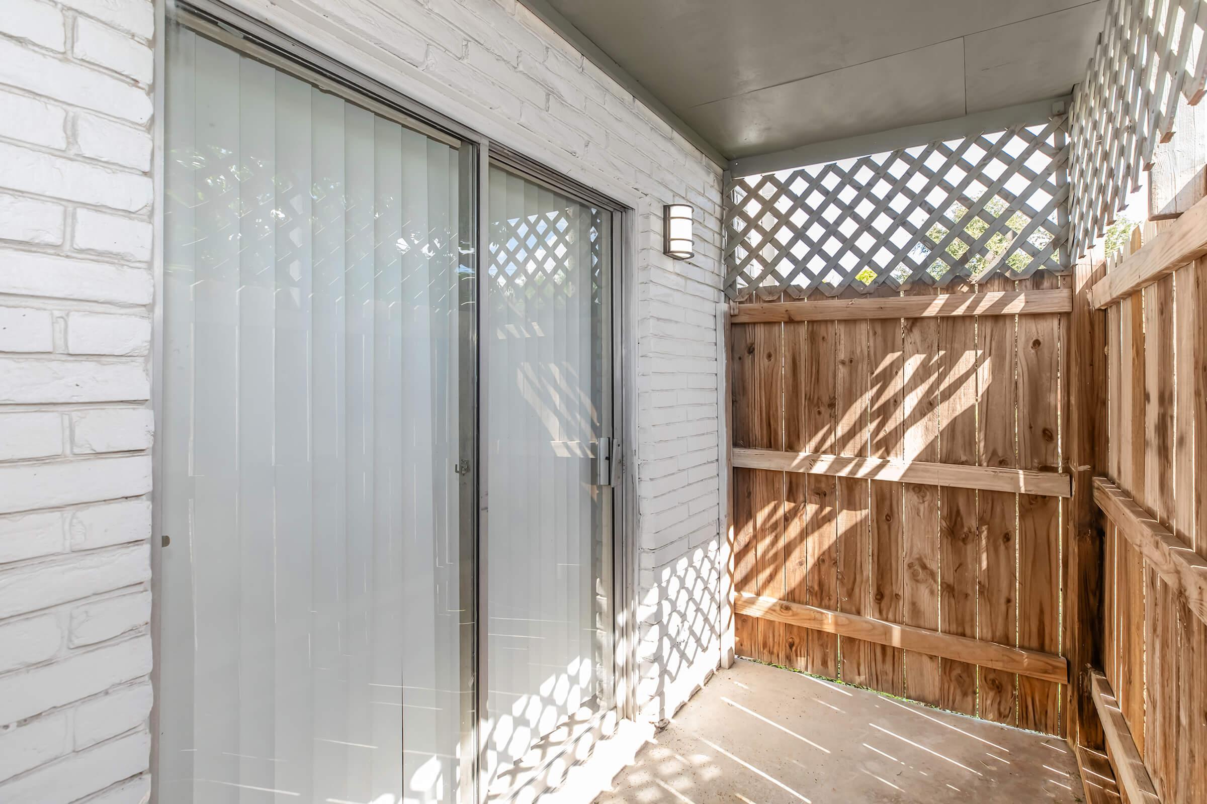 A small outdoor space featuring a wooden fence, vertical blinds on glass sliding doors, and a lattice overhead. Sunlight casts shadows on the ground, creating a cozy and private atmosphere.