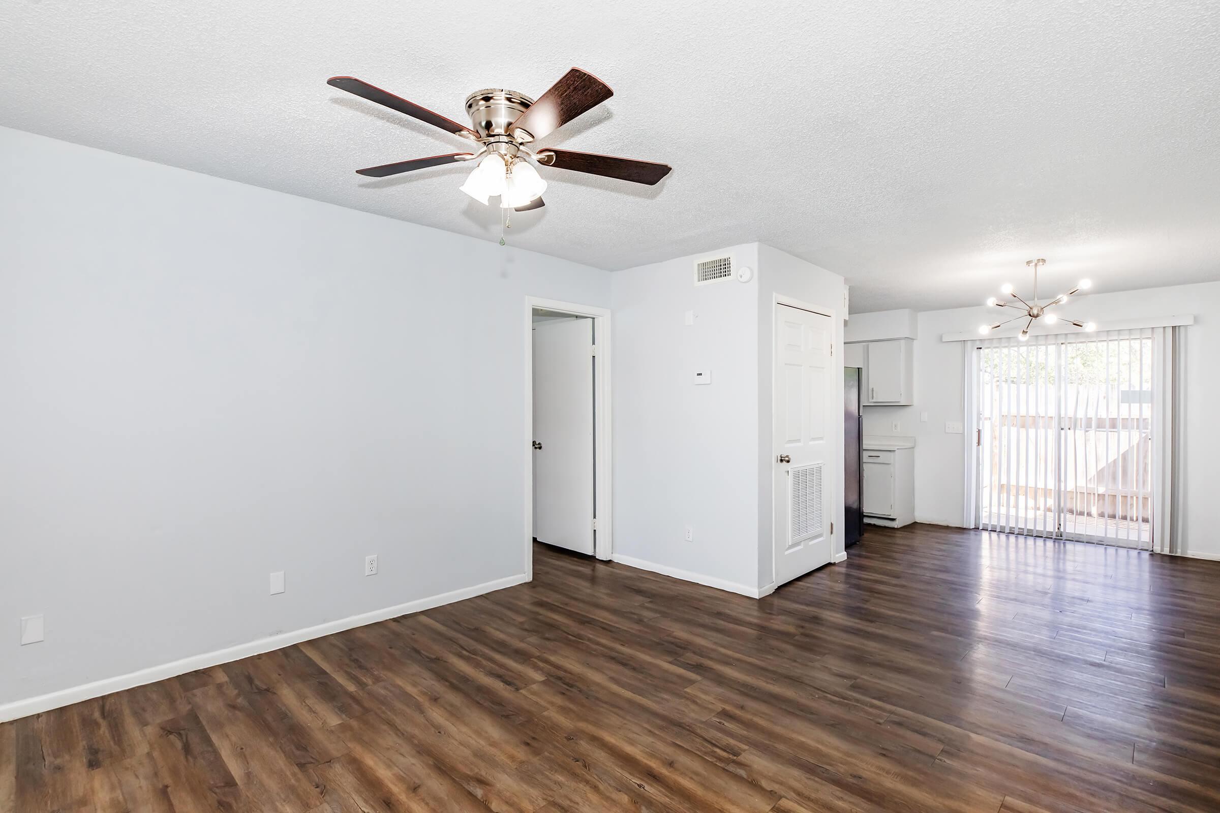 Spacious living area with a ceiling fan and modern light fixtures, featuring light gray walls and a wood-style floor. A doorway leads to a kitchenette, and sliding glass doors open to an outdoor area, providing natural light. The overall design is bright and inviting.