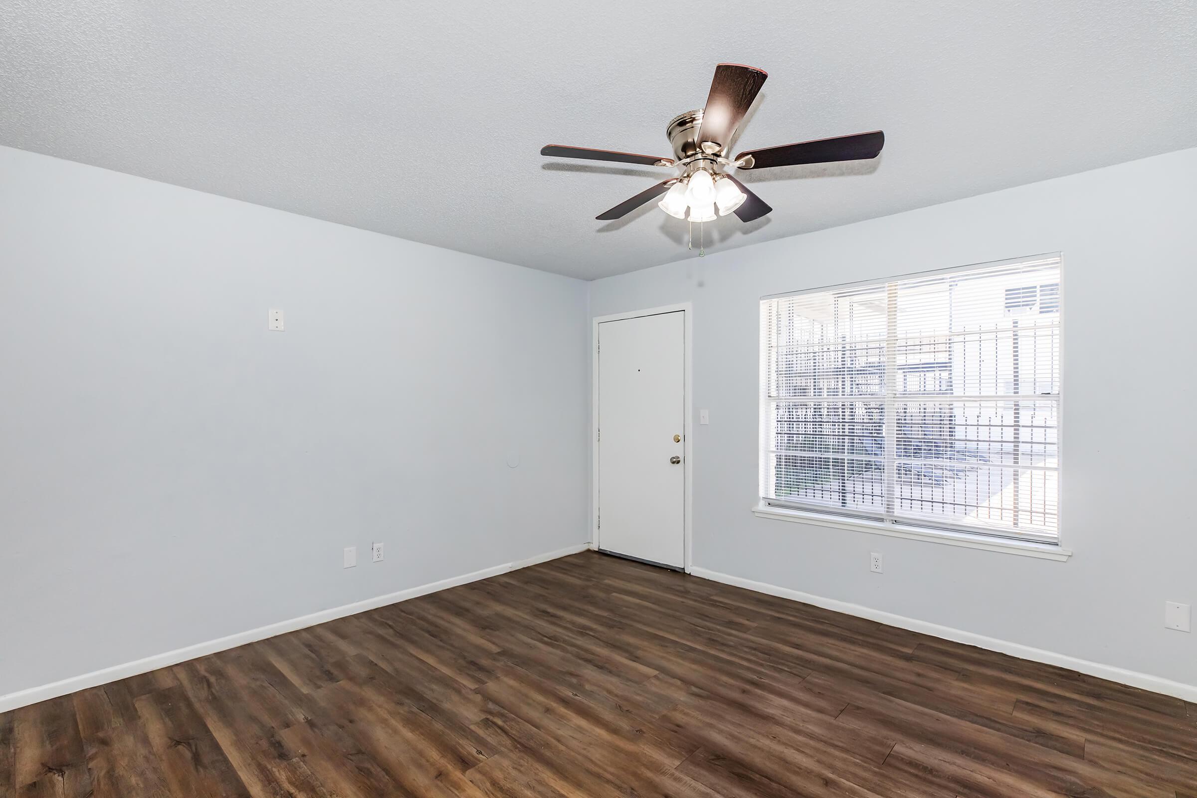 A clean, empty living room with light blue walls and brown wooden flooring. There's a ceiling fan with light fixtures, and a window showing outdoor light. The door is located on the left side of the room, and natural light illuminates the space.