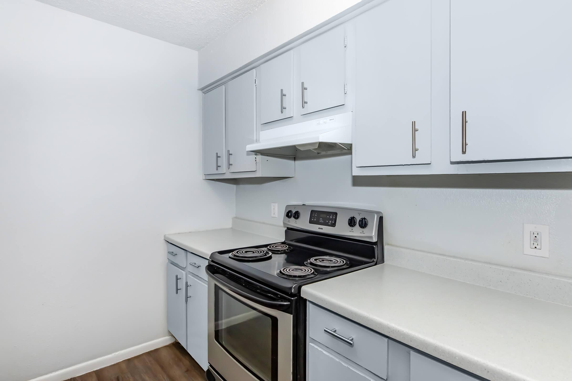 Modern kitchen with light blue cabinets, a stainless steel oven and stove, and white countertops. The walls are painted a soft gray, and the flooring is a wood-like laminate. There is ample cabinet space above and a range hood above the stove.
